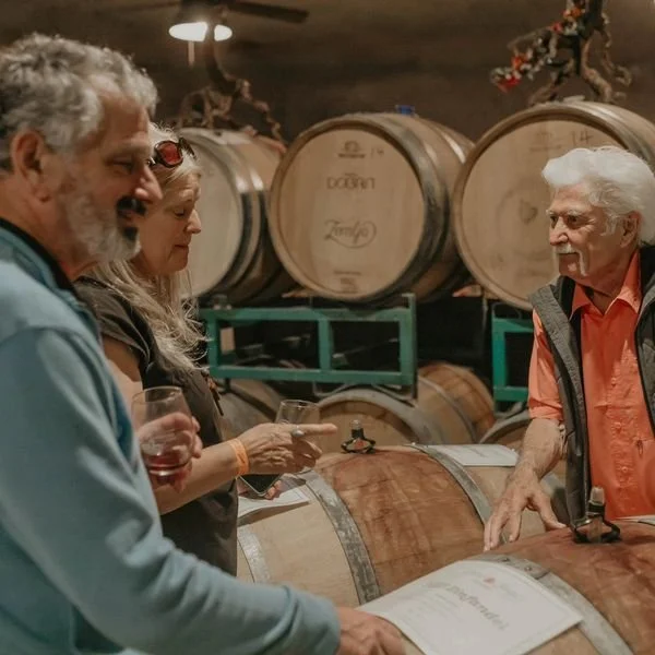 Three people tasting wine and looking at a menu inside a winery, with large wooden wine barrels in the background.