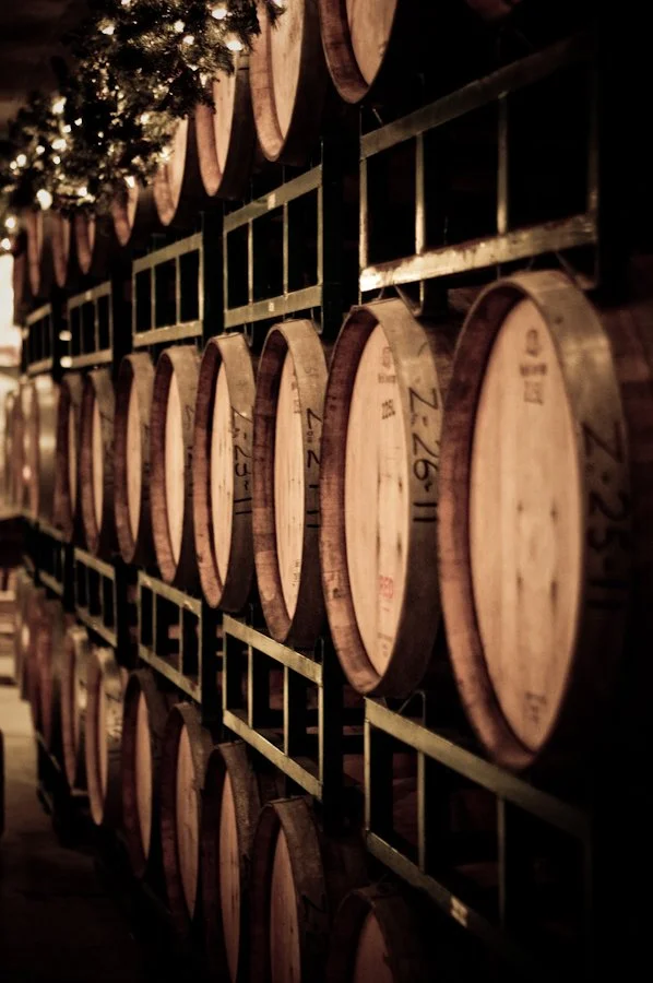 Rows of wooden wine barrels stacked on metal racks in a cellar, with warm lighting and a decorated green garland with Christmas lights at the top.