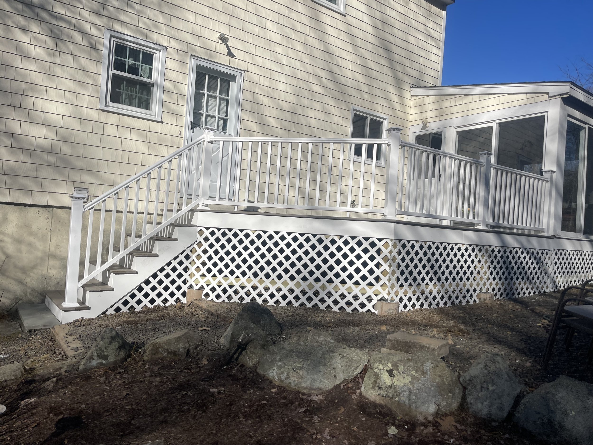 White wooden deck with stairs leading up to a door on the back of a house, with a lattice skirting underneath and windows on the house's exterior wall.