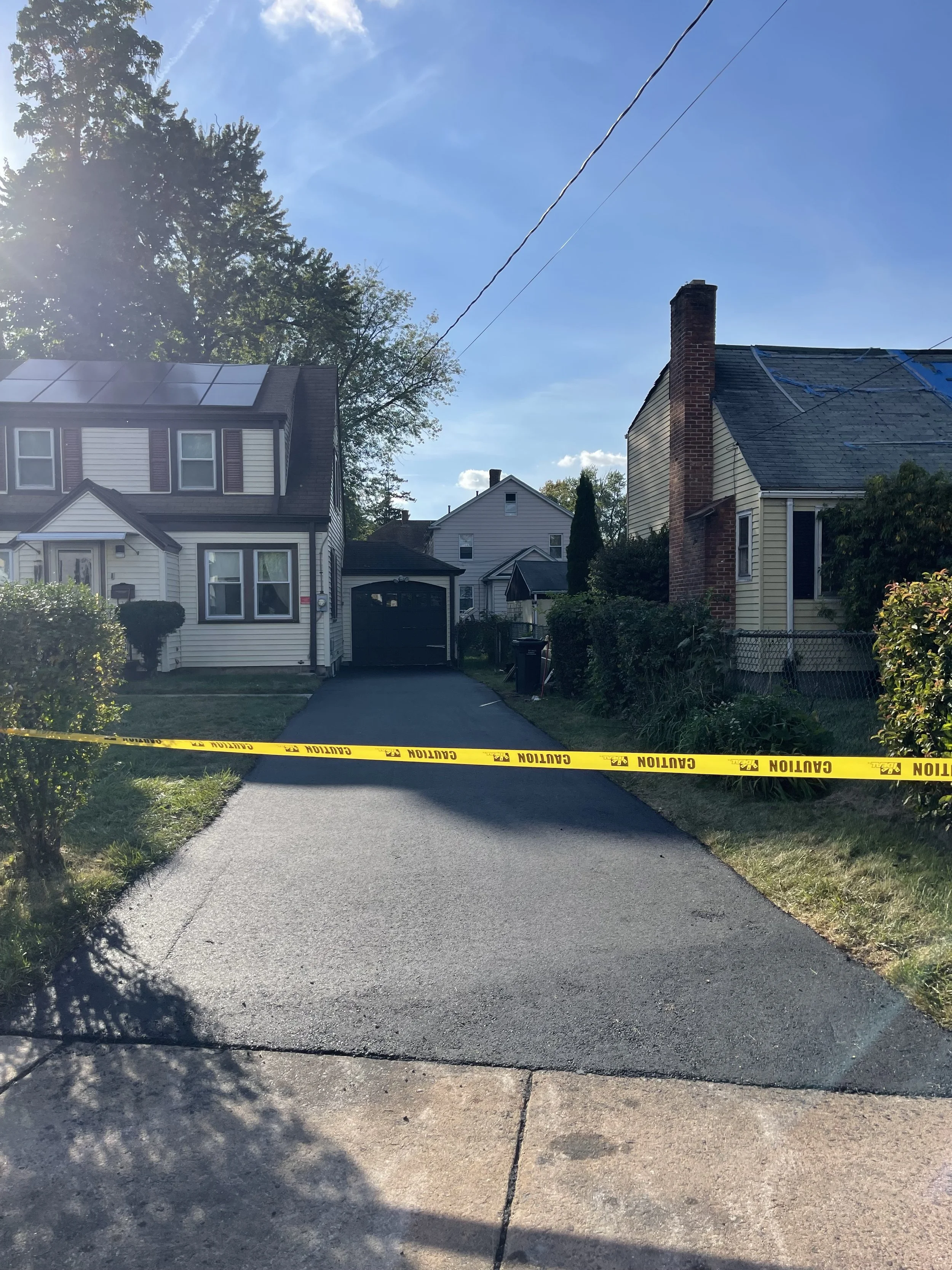 Residential driveway blocked with yellow caution tape, houses on either side, clear sky, and sunlight casting shadows.