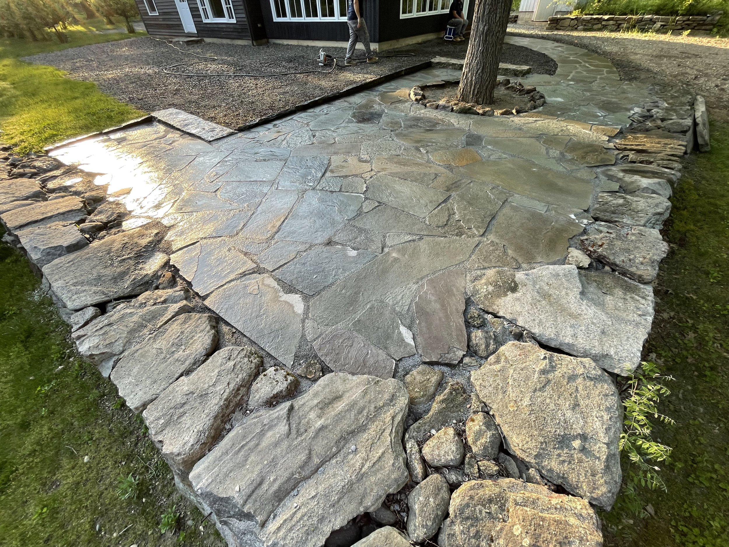 A newly constructed stone patio with large flat stones and a border of larger, rougher stones surrounds a tree in a backyard. A person is working nearby, and a house is visible in the background.