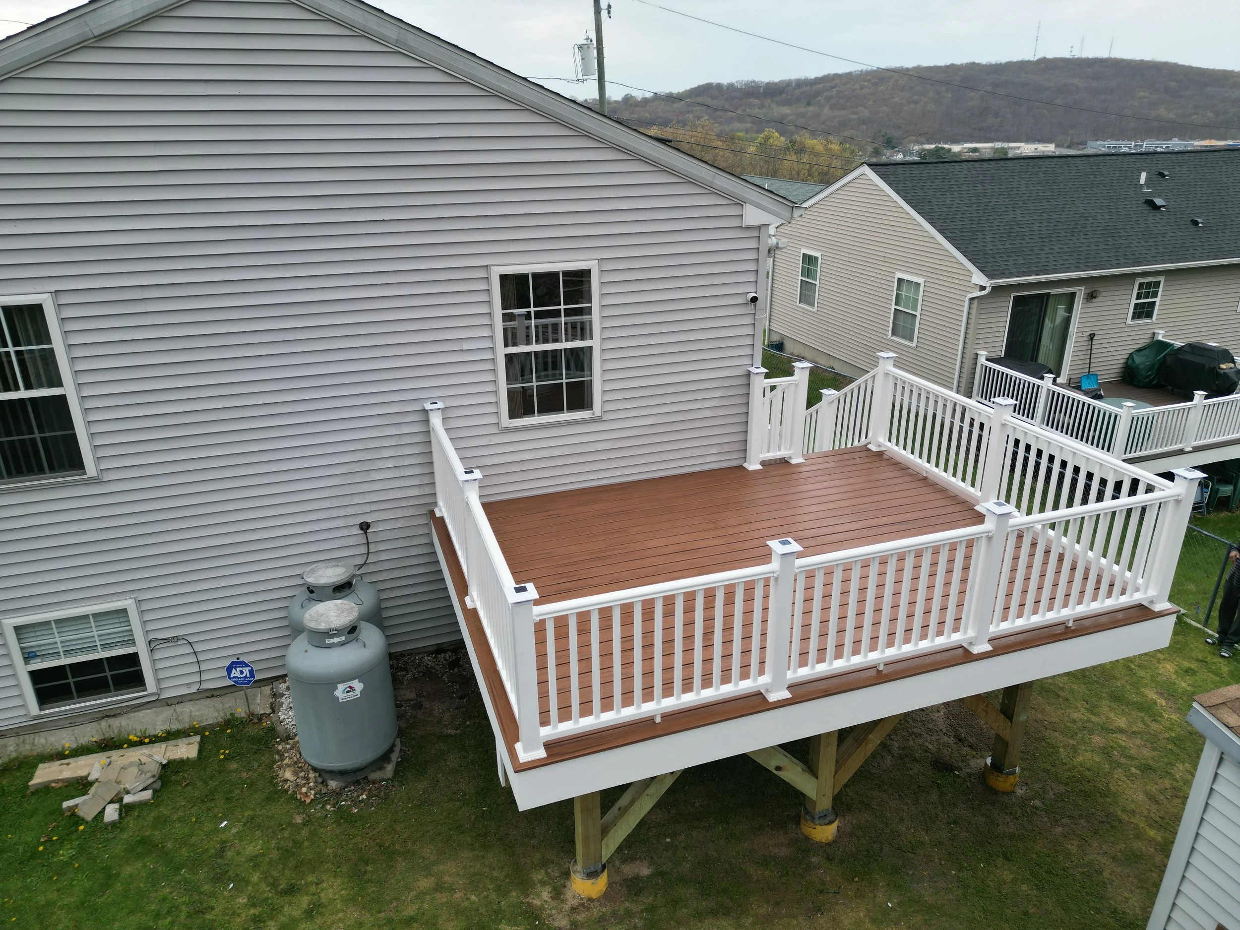 An outdoor wooden deck attached to a house with beige siding, white railing, and steps leading down. Two gas tanks and an ADT security sign are near the house. In the background, there are neighboring houses and rolling hills.