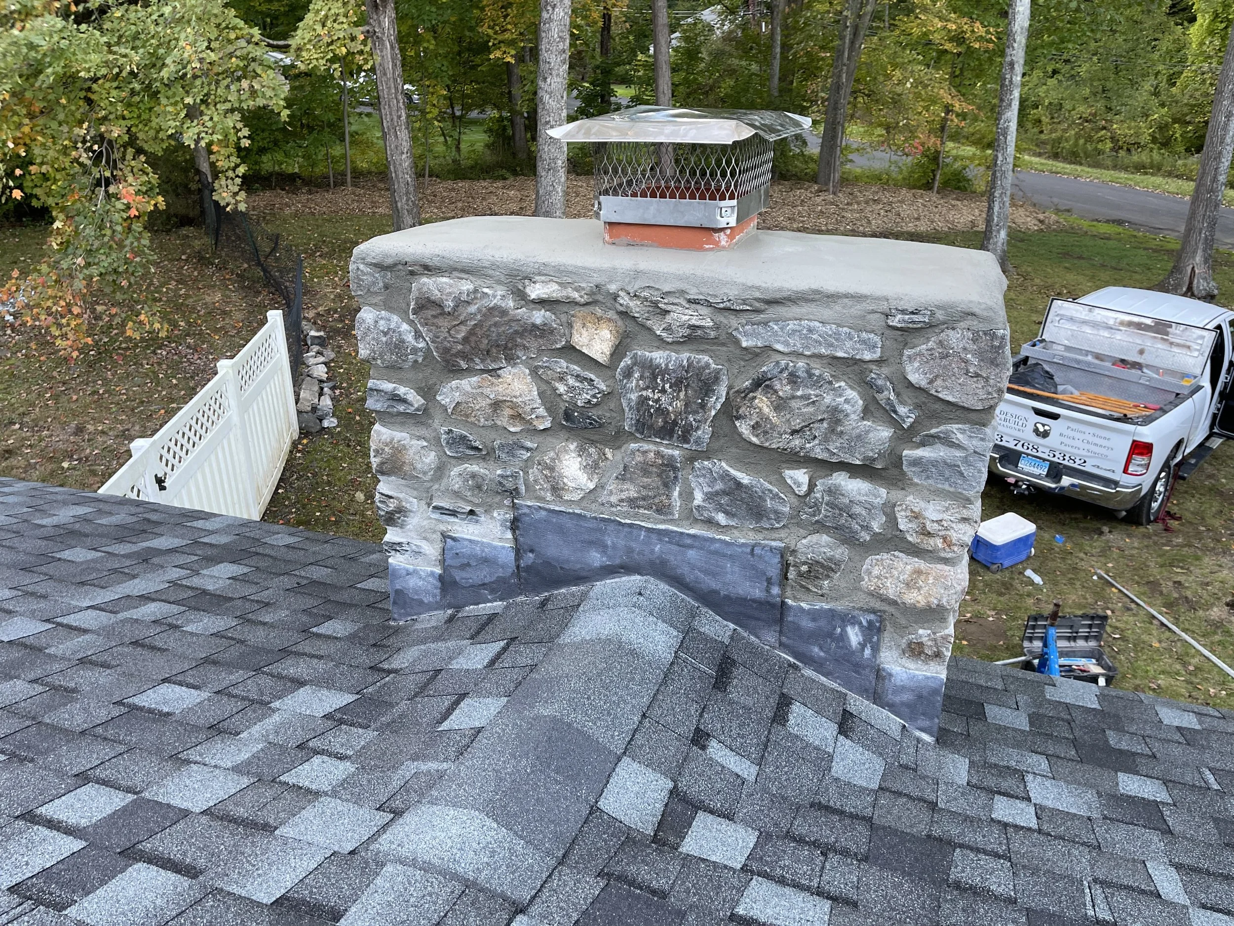 View of a stone chimney on a roof, with a metal vent cap on top, surrounded by trees and a driveway with a white truck parked nearby.