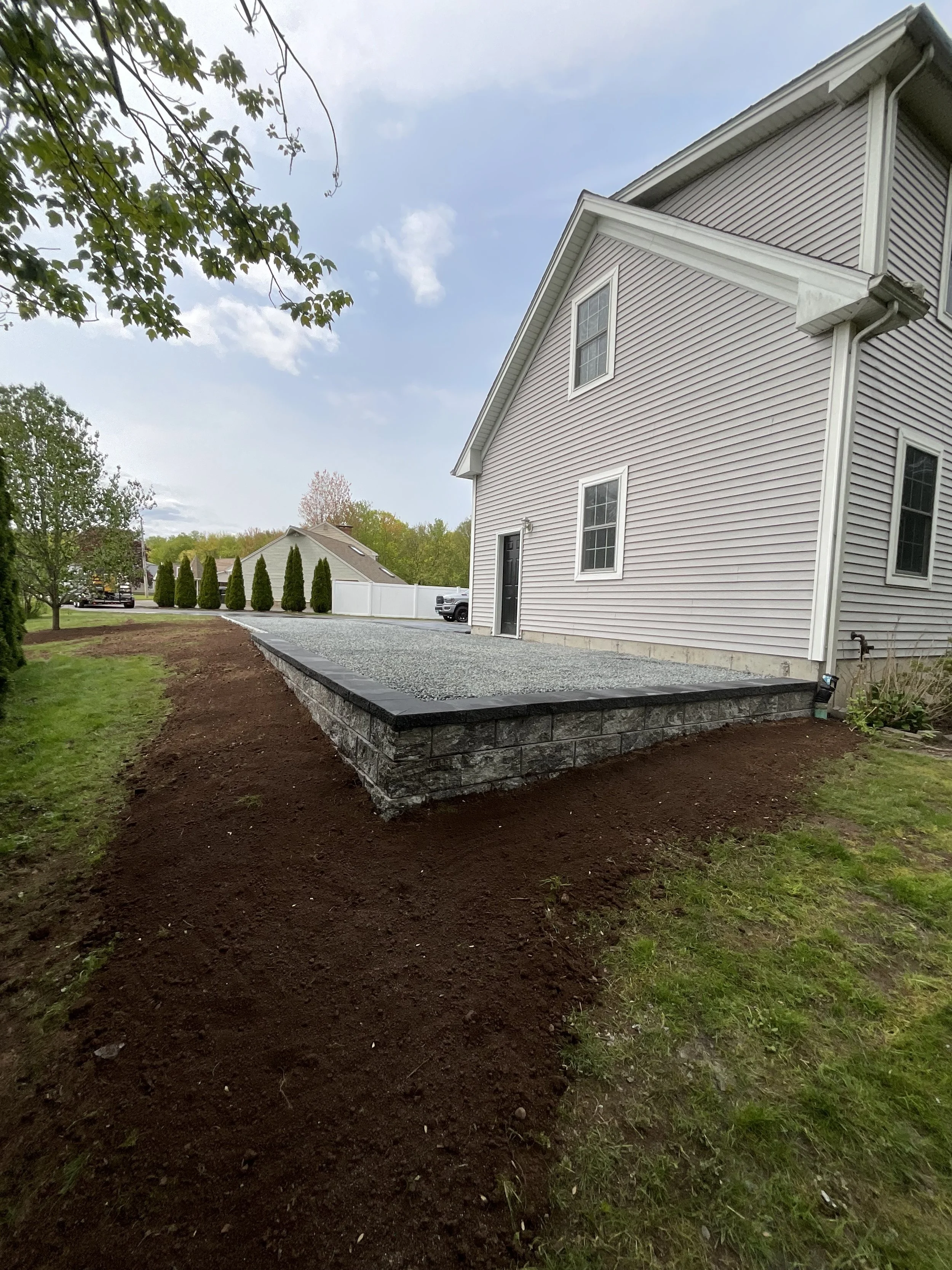 A newly built raised stone and gravel patio or deck beside a light pink house with white trim. The patio has a black stone edge and is surrounded by freshly tilled soil, with a grass lawn nearby. There are trees in the background under a partly cloud