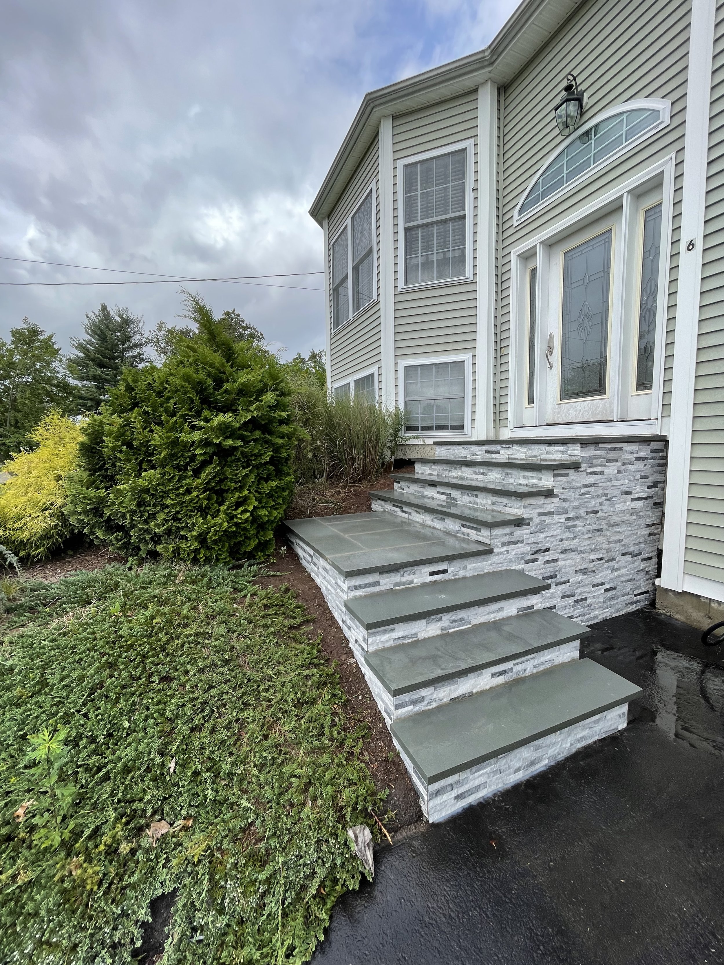 Exterior view of a house showing a front porch with grey and white stone stairs, green siding, and a white front door with stained glass, surrounded by landscaping and a wet black asphalt driveway.