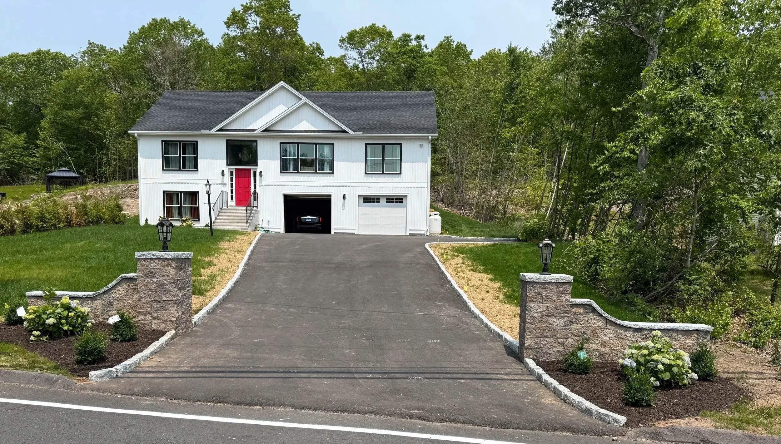 A white two-story house with black roof and a red front door, surrounded by green lawns and trees, with a paved driveway and stone pillars with lamps at the entrance.