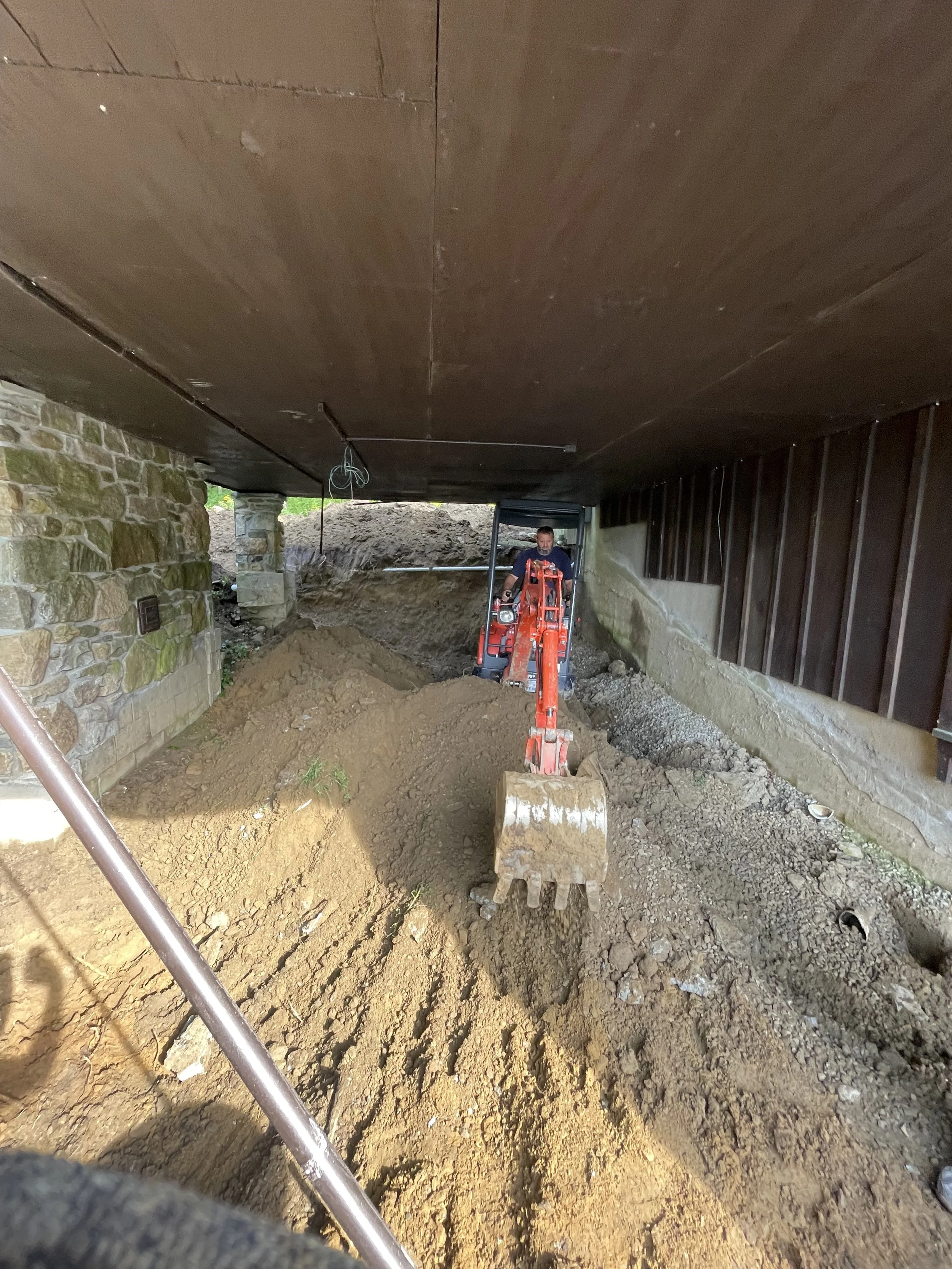 Construction worker operating a small excavator underground, moving dirt beneath a building with stone and wood walls.