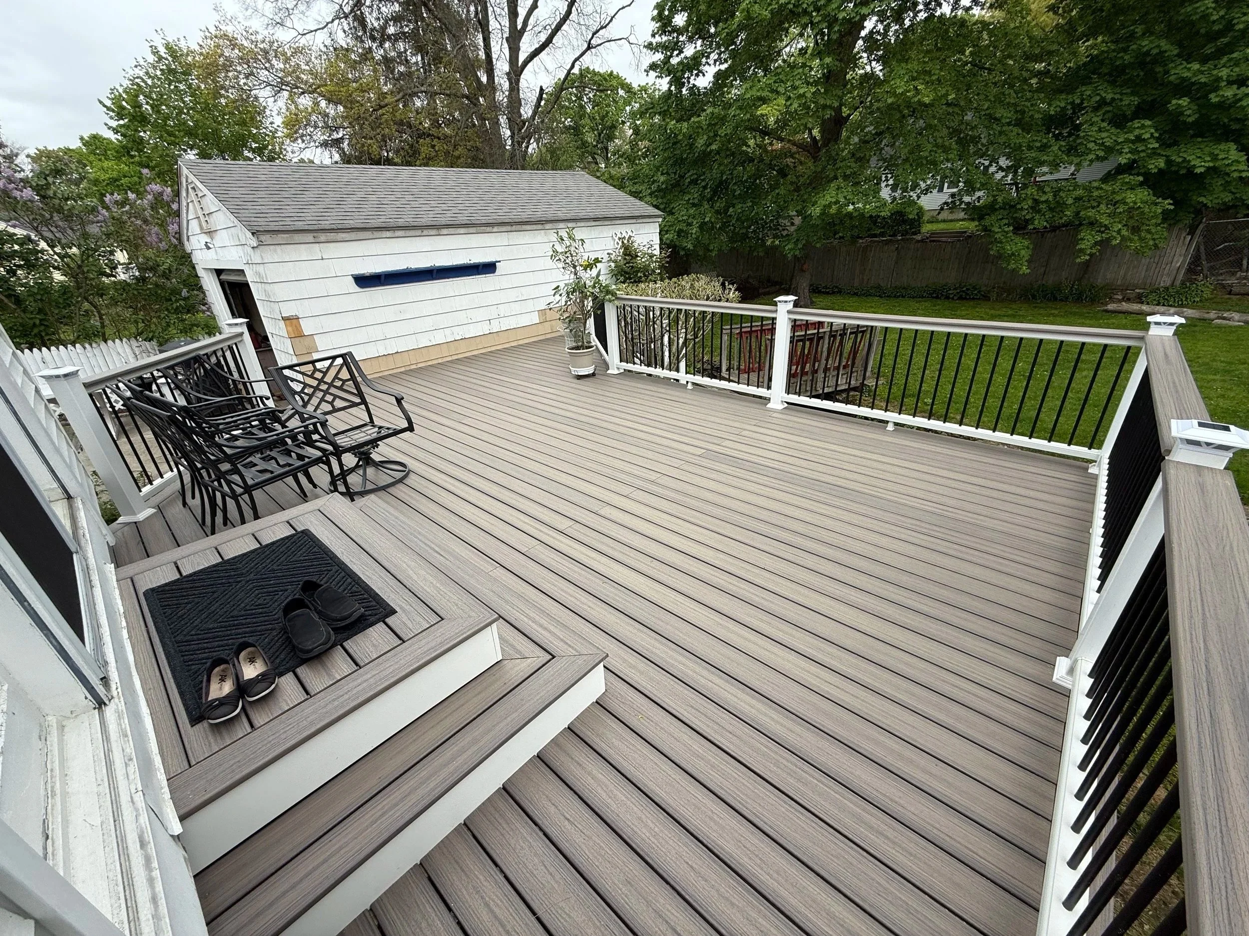 A spacious outdoor deck with light brown wooden flooring, black metal chairs on the left, a white staircase with a black rubber mat and shoes at the base, and a white railing with black balusters. In the background, there is a white shed with a gray shingle roof, a potted plant, and lush green trees and grass.