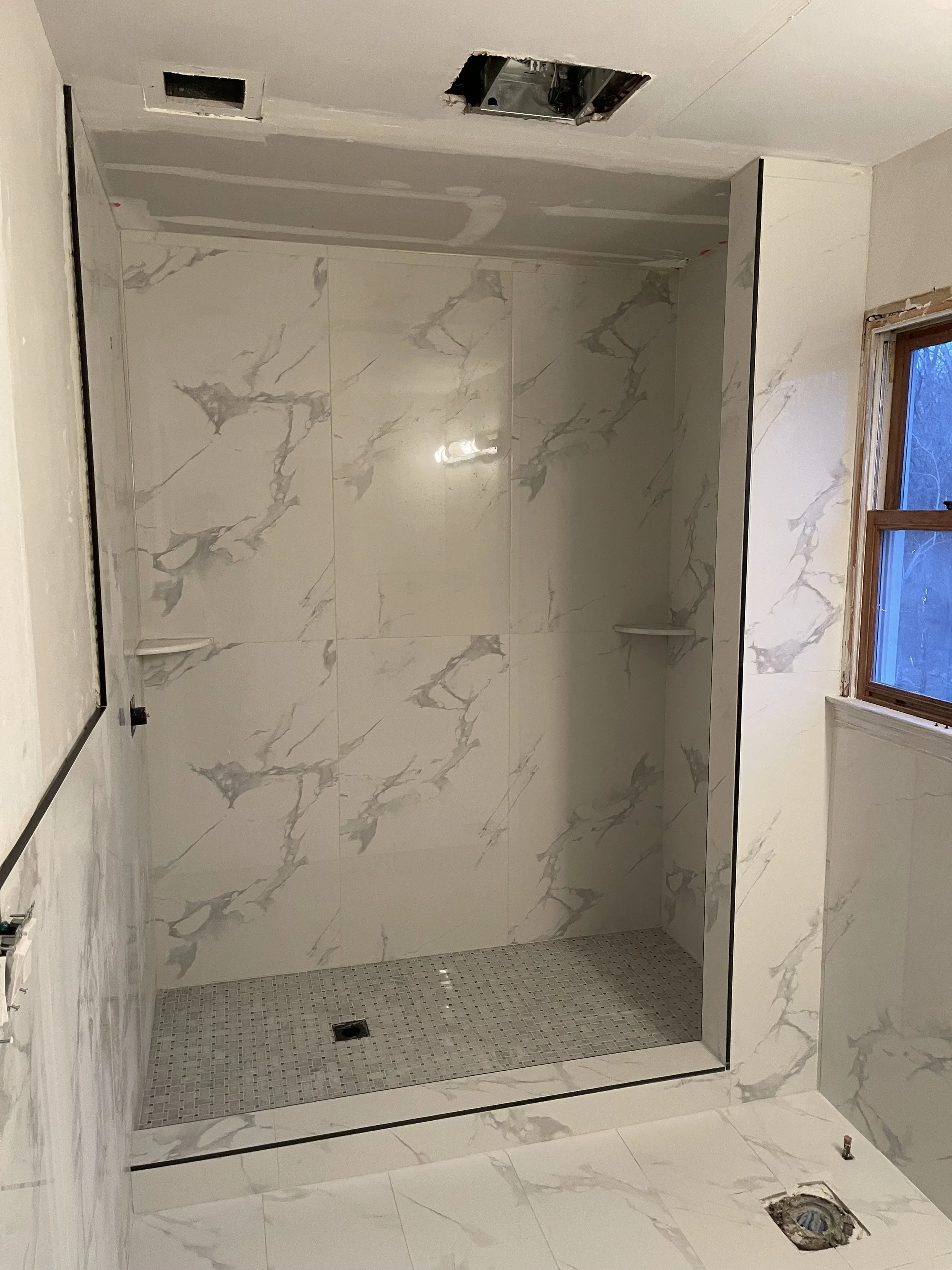 unfinished bathroom shower stall with marble tile walls and floor, window on right, and exposed plumbing and ceiling fixtures.