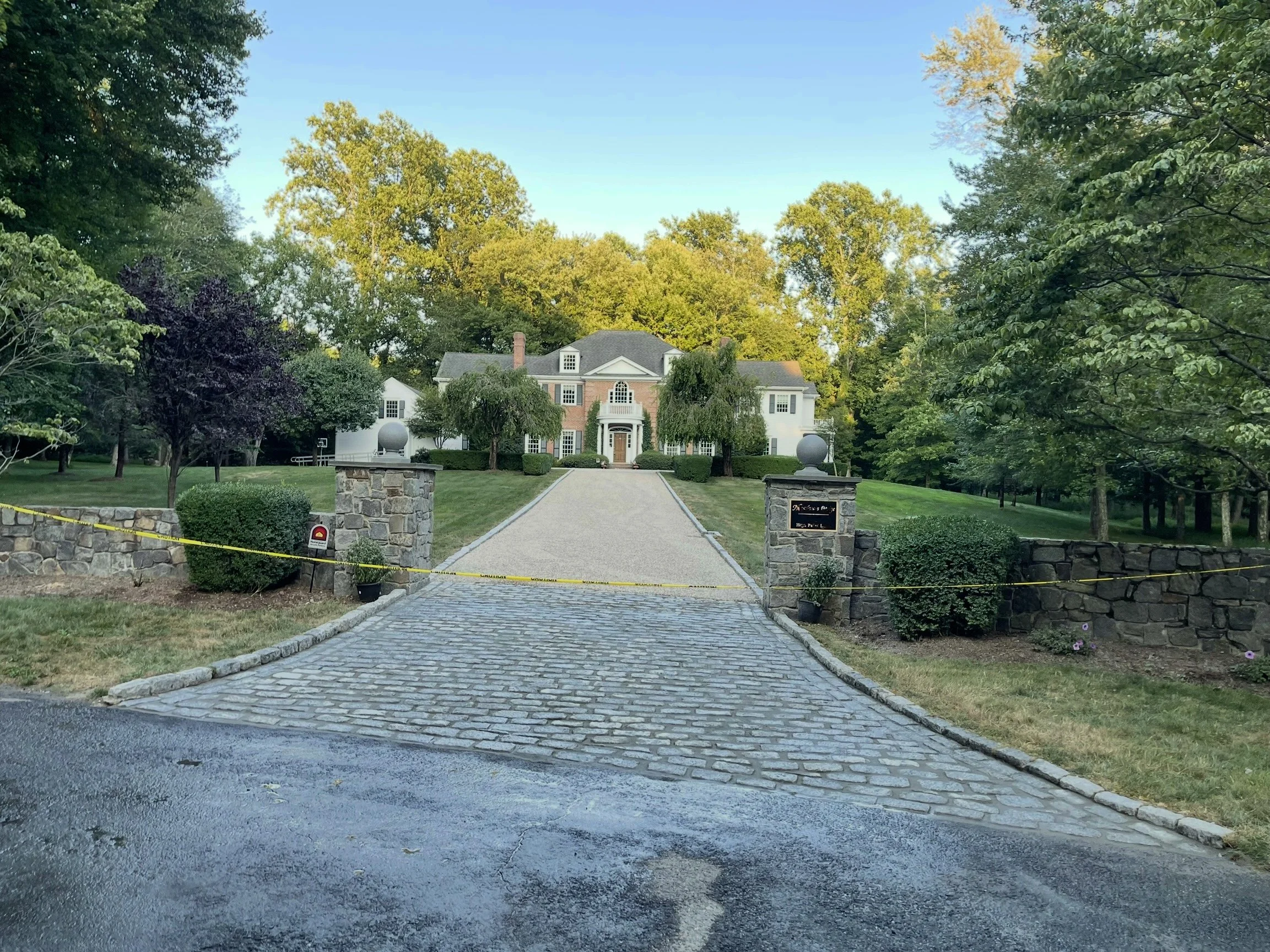 Large white house with brick accents and a gravel driveway, flanked by stone pillars and lush green trees, with a black and yellow caution tape across the driveway.
