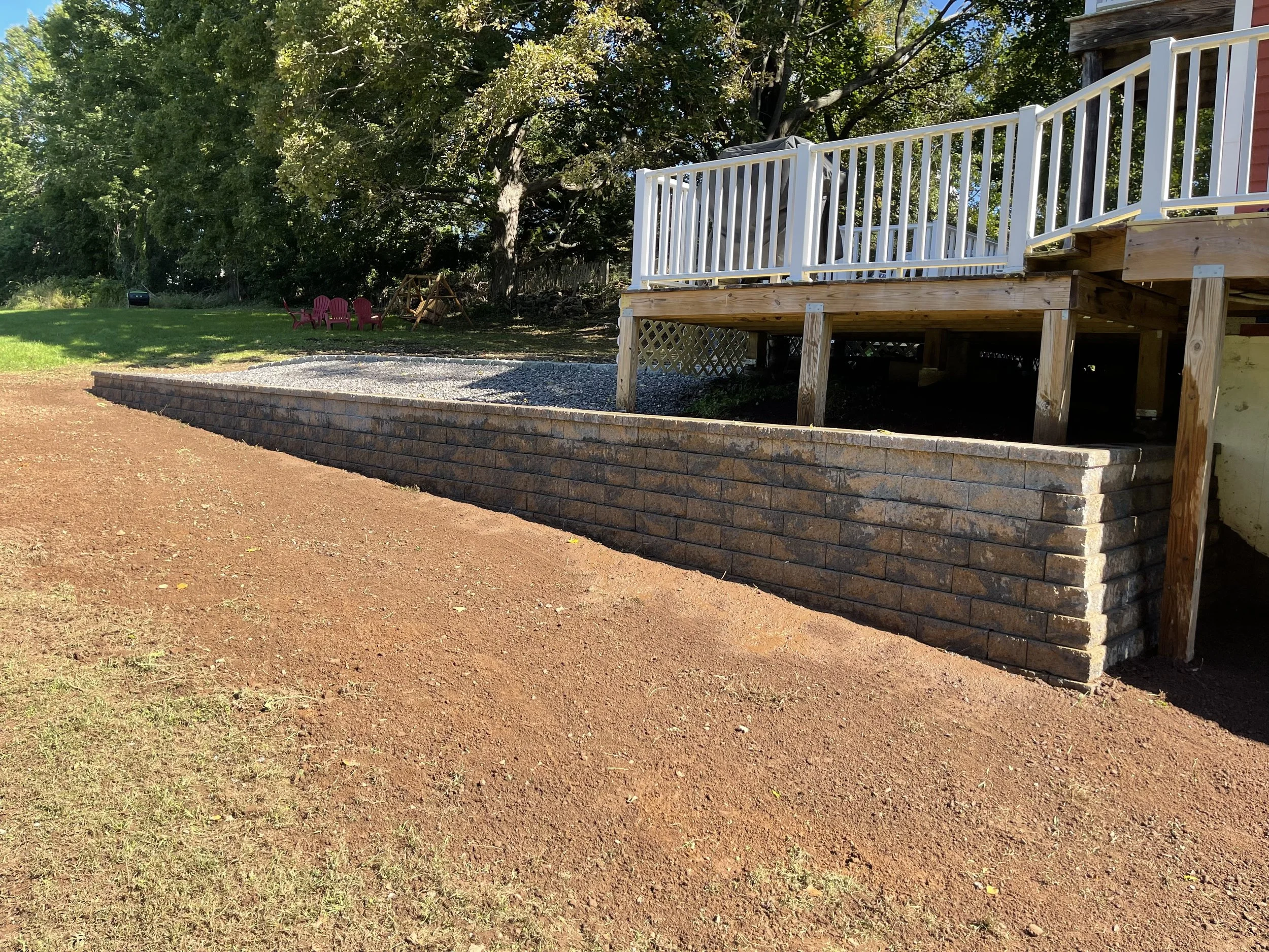 Backyard with a new brick retaining wall, a wooden deck with white railing, and a grassy area with trees and pink outdoor chairs.