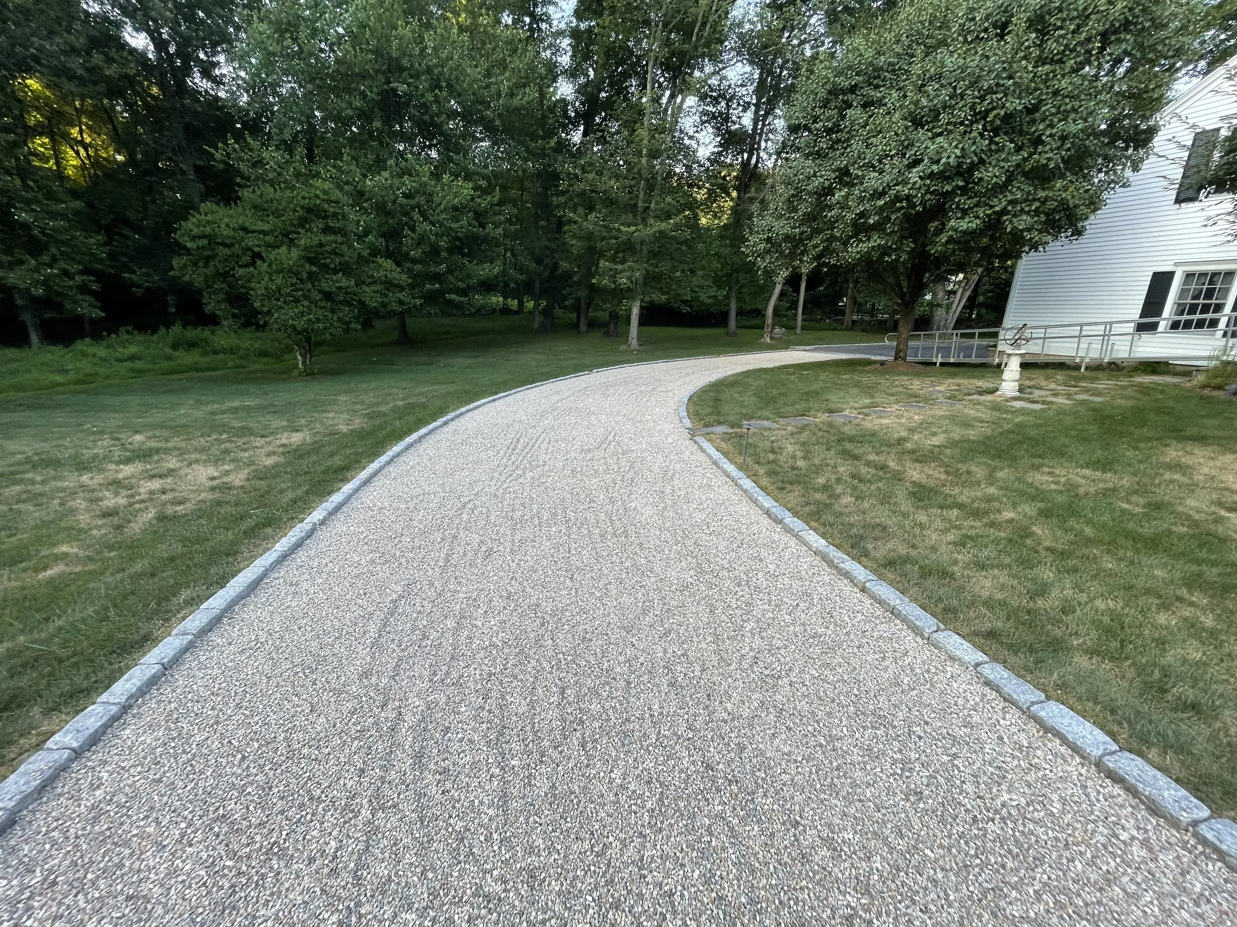 Gravel pathway curving through a landscaped yard with grass, trees, and a white house with a ramp.