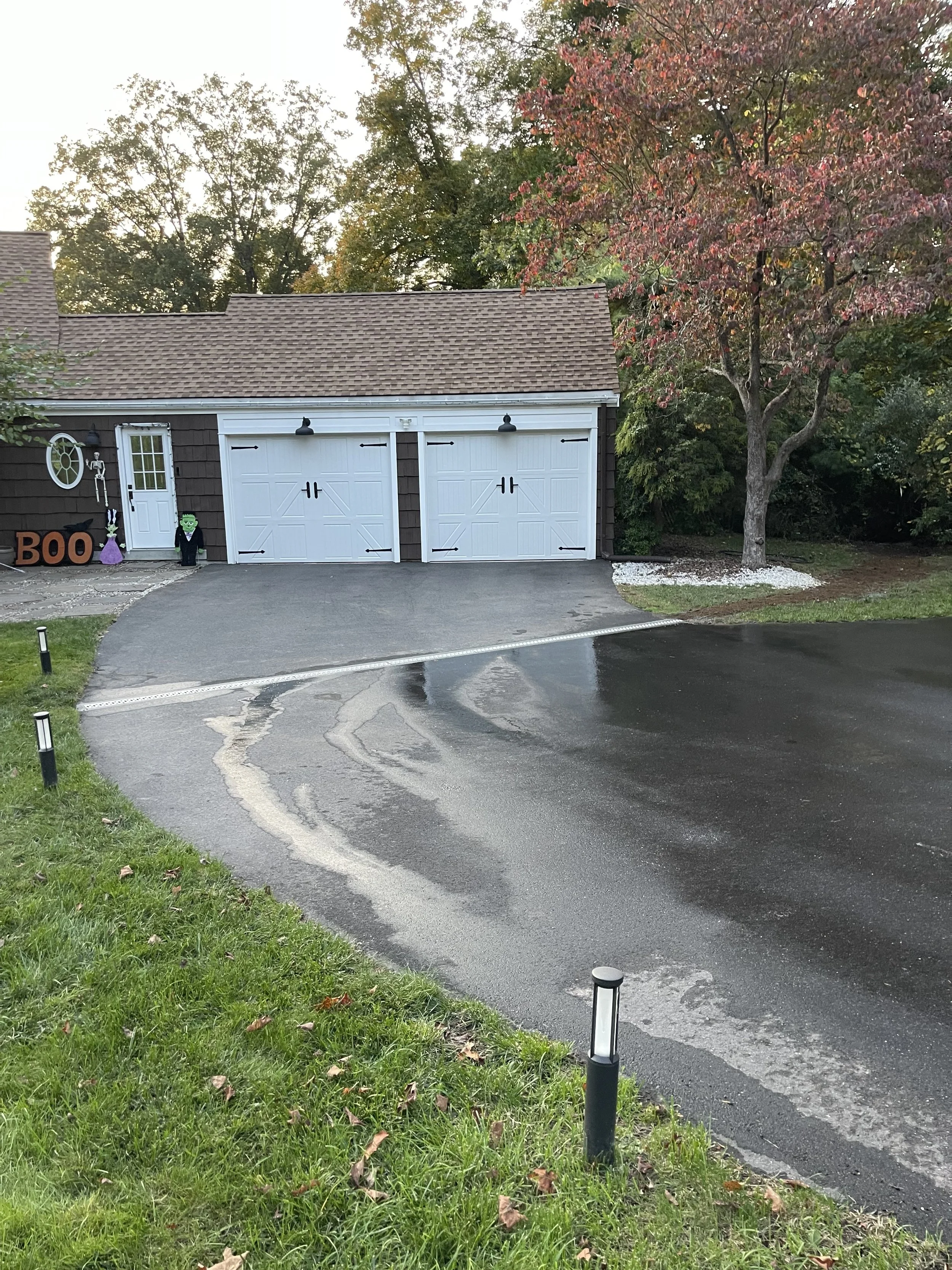 A residential garage with Halloween decorations, including a ghost, skeleton, witch, and the word 'BOO' in orange letters. The driveway is wet, reflecting the sky, and surrounded by green grass and small black pathway lights. A tree with red and green leaves is on the right side.