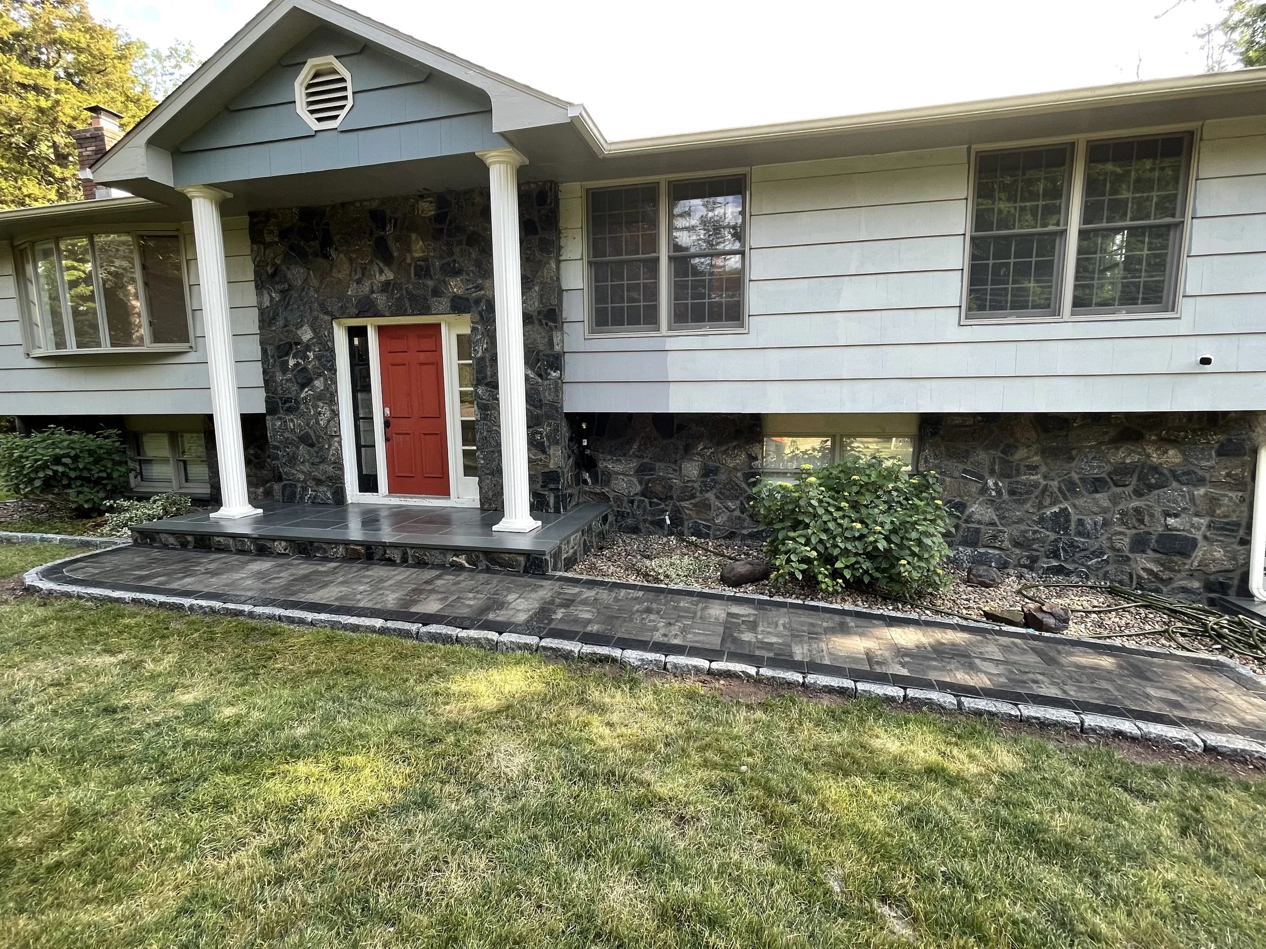 Front view of a house with a red door, white columns, and a stone and siding exterior. There is a paved walkway and lawn in the foreground.