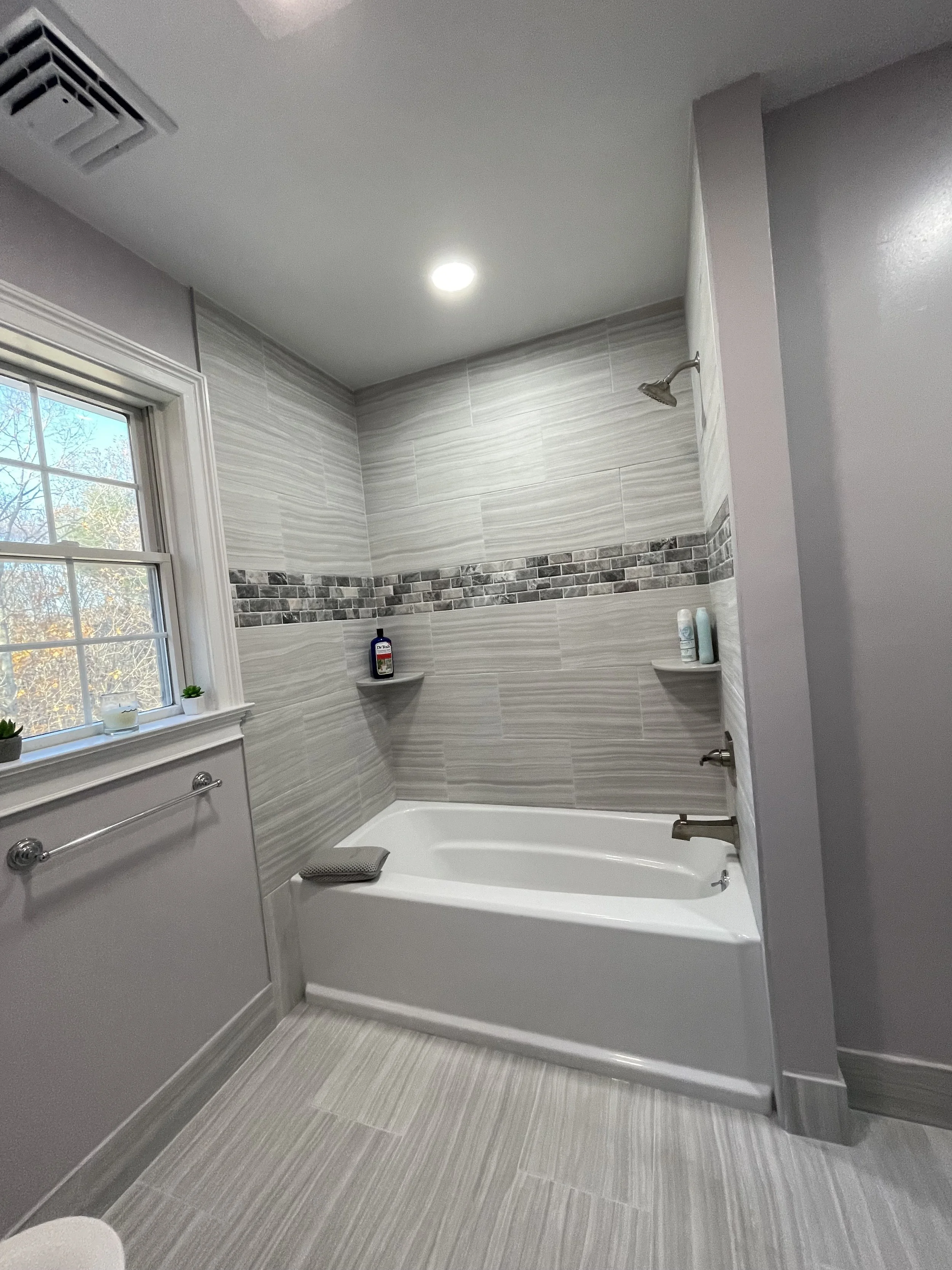 Bathroom with a bathtub, gray tiled walls with a decorative strip, window with plants, and a shelf with toiletries.