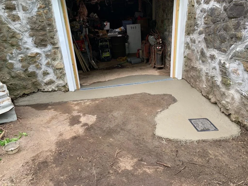 A stone wall with a newly poured concrete or cement slab in front of a partially open garage door, with some gardening tools and stored items visible inside.