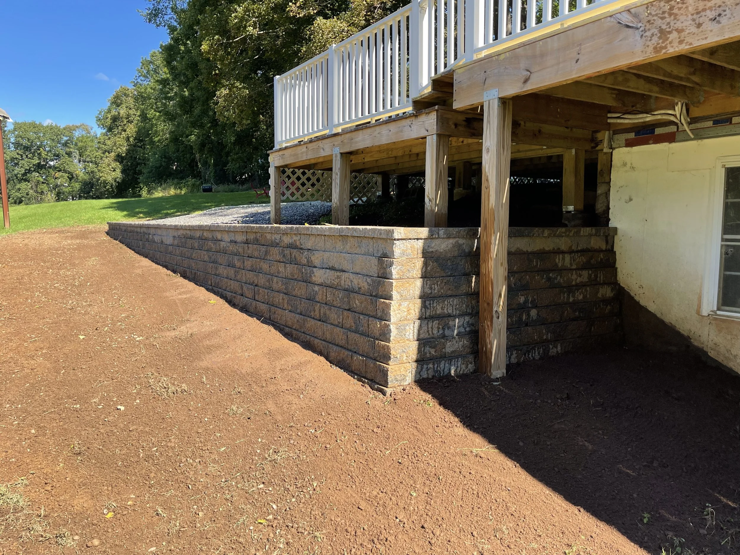 A brick retaining wall under a wooden deck with stairs leading down to the ground, beside a house with a window and doorway, on a sunny day with a blue sky.