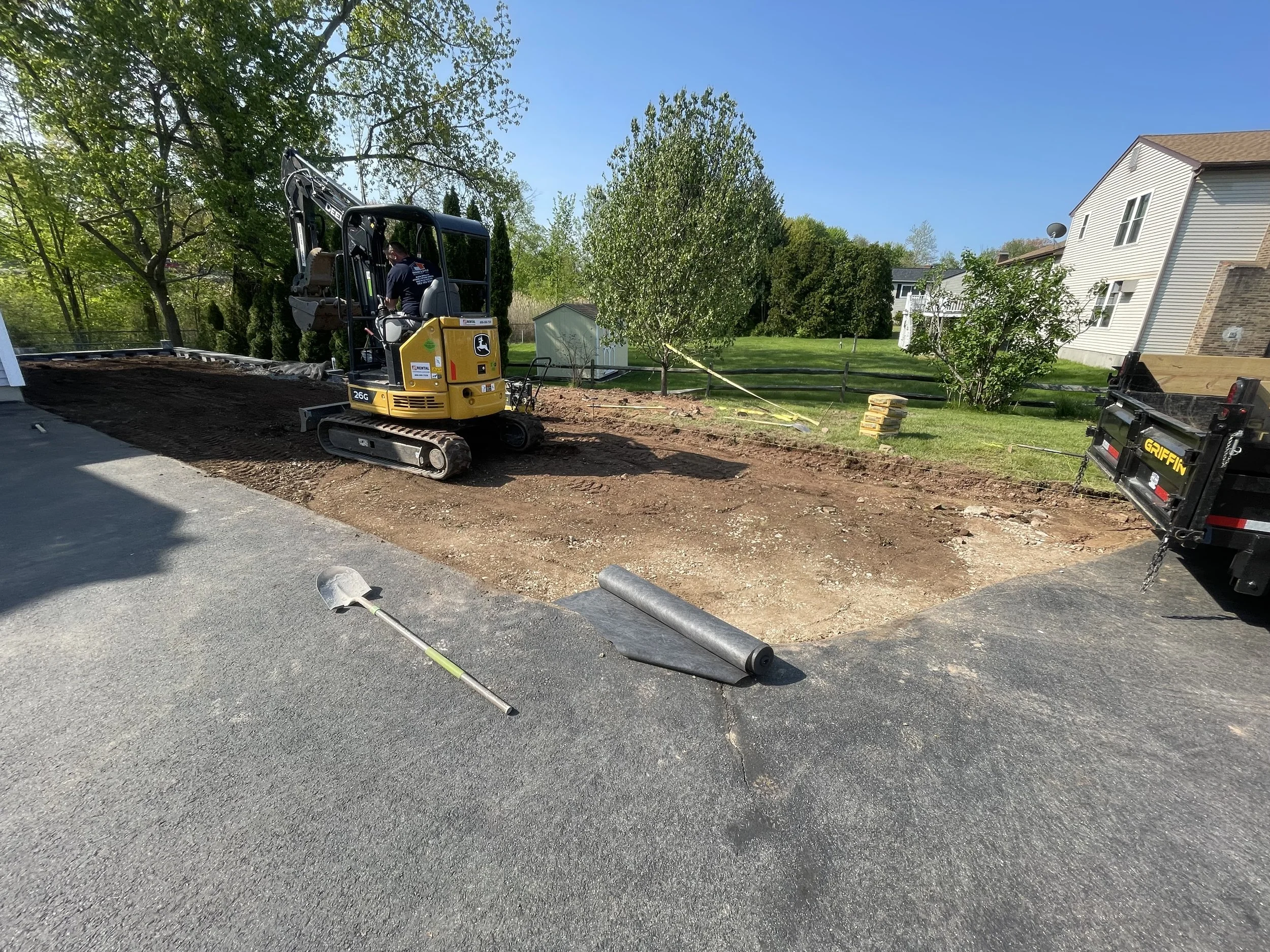 Construction site with a mini excavator digging on a residential property, with a wheelbarrow and rolled landscaping fabric near the edge of the excavation, and a black dump truck with yellow accents to the right, surrounded by green trees, grass, and neighboring houses under a clear blue sky.