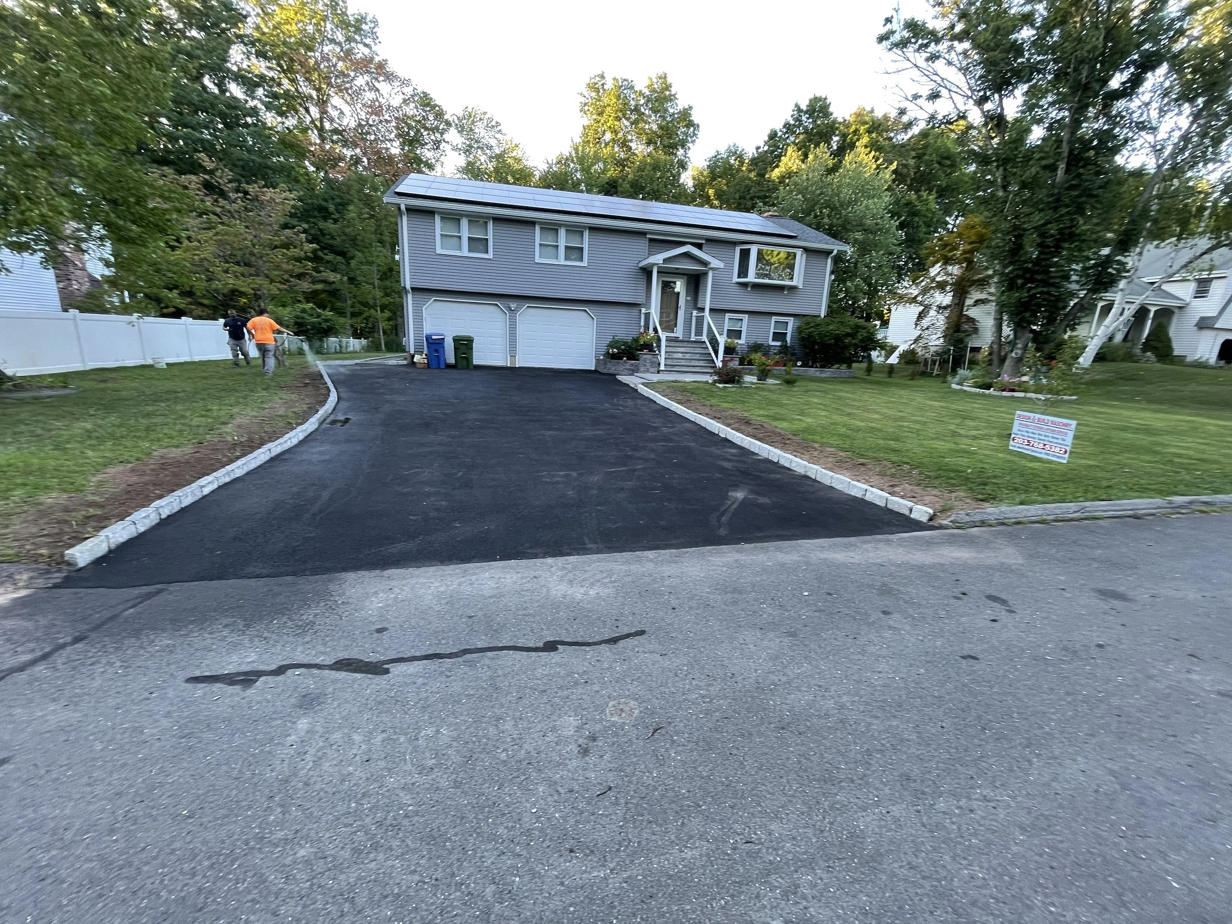 Freshly paved black asphalt driveway leading up to a two-story gray house with a white garage door and solar panels on the roof. The driveway is bordered by white stone curbs and green lawns with trees and bushes. Two workers are walking away on the left side. A sign on the lawn indicates driveway work.