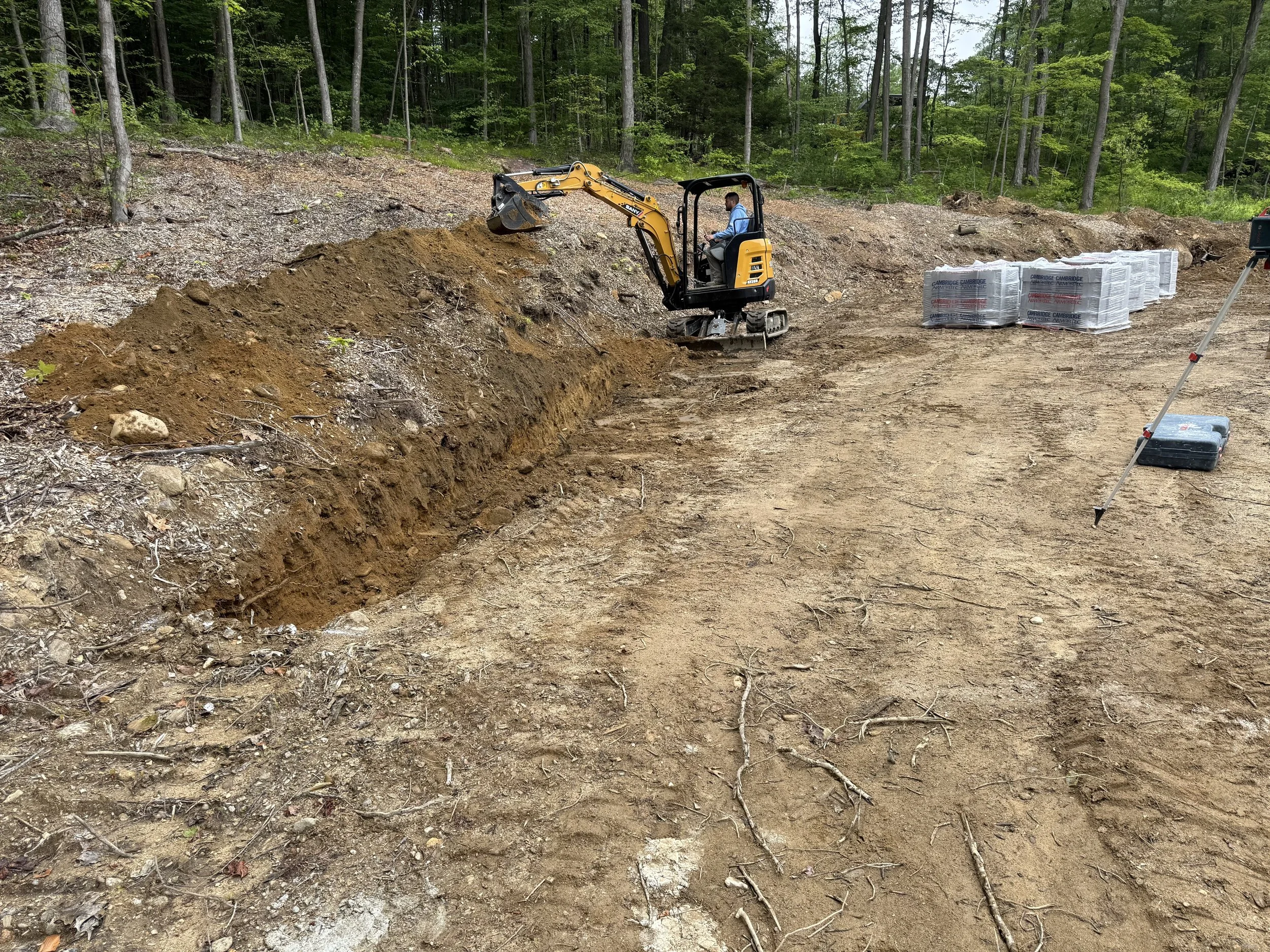 A person operating a small excavator on a construction site, with a trench being dug into the dirt, and stacks of building materials in the background amid a wooded area.