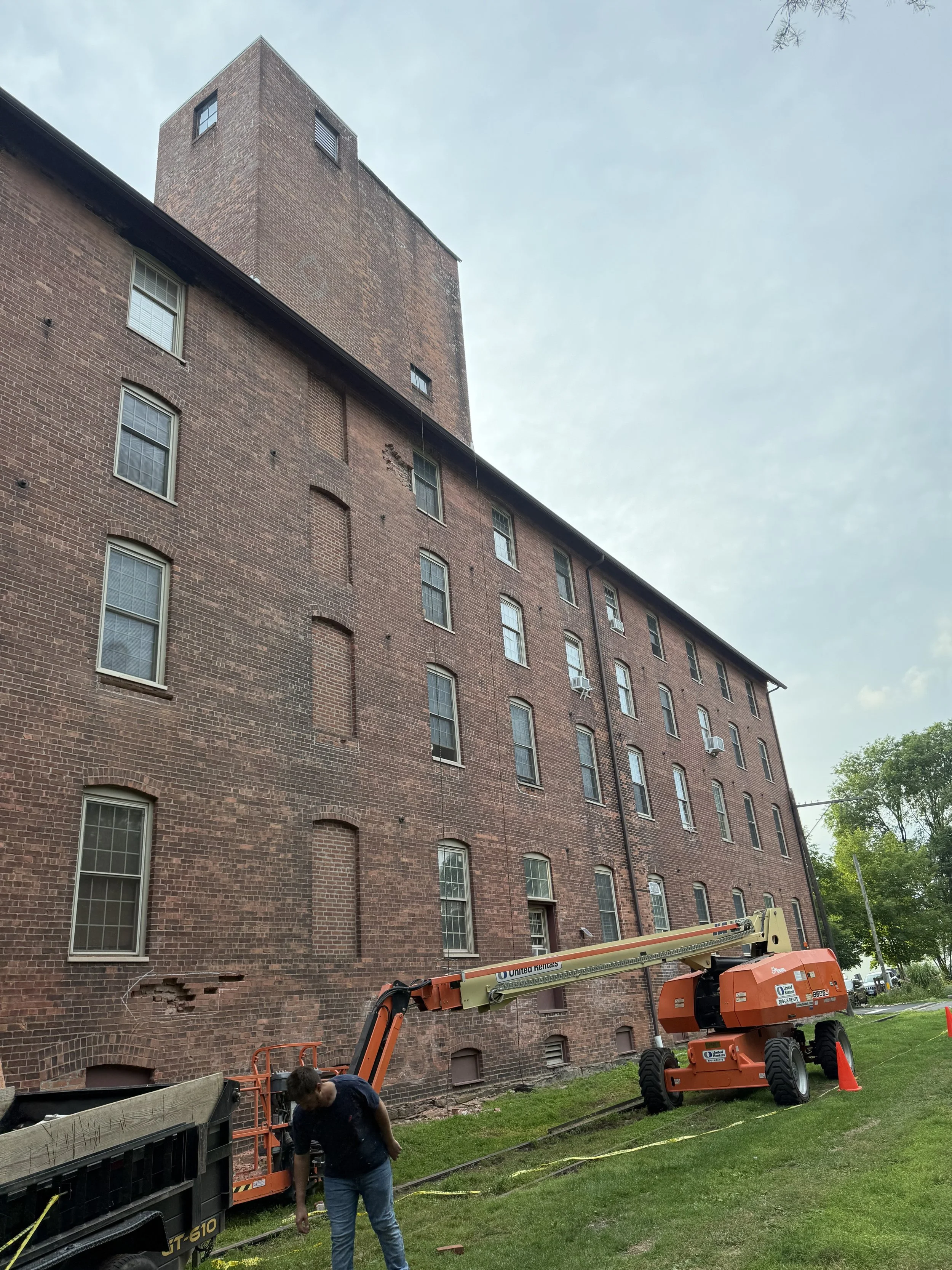 A man working near a cherry picker lift beside a large brick building with multiple windows and a tower on top, on a cloudy day.