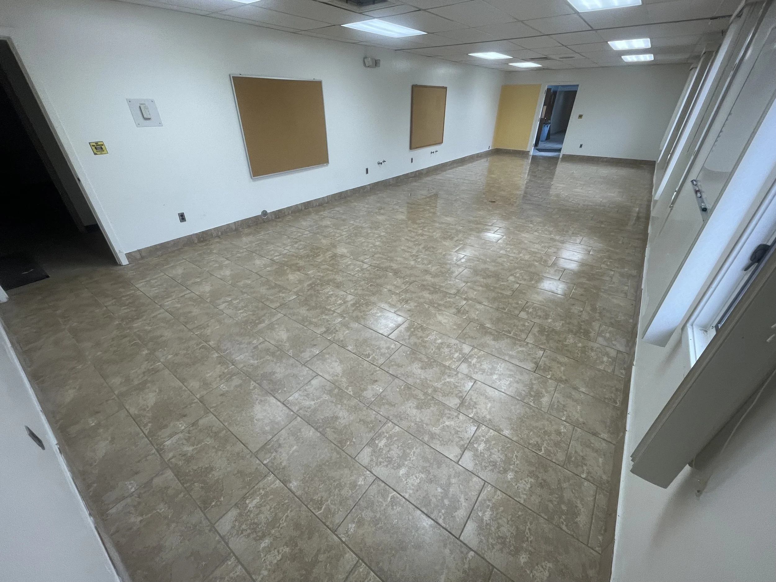 Empty room with beige tiled floor, white walls, cork bulletin boards, and fluorescent ceiling lights. There is a doorway leading to another room.