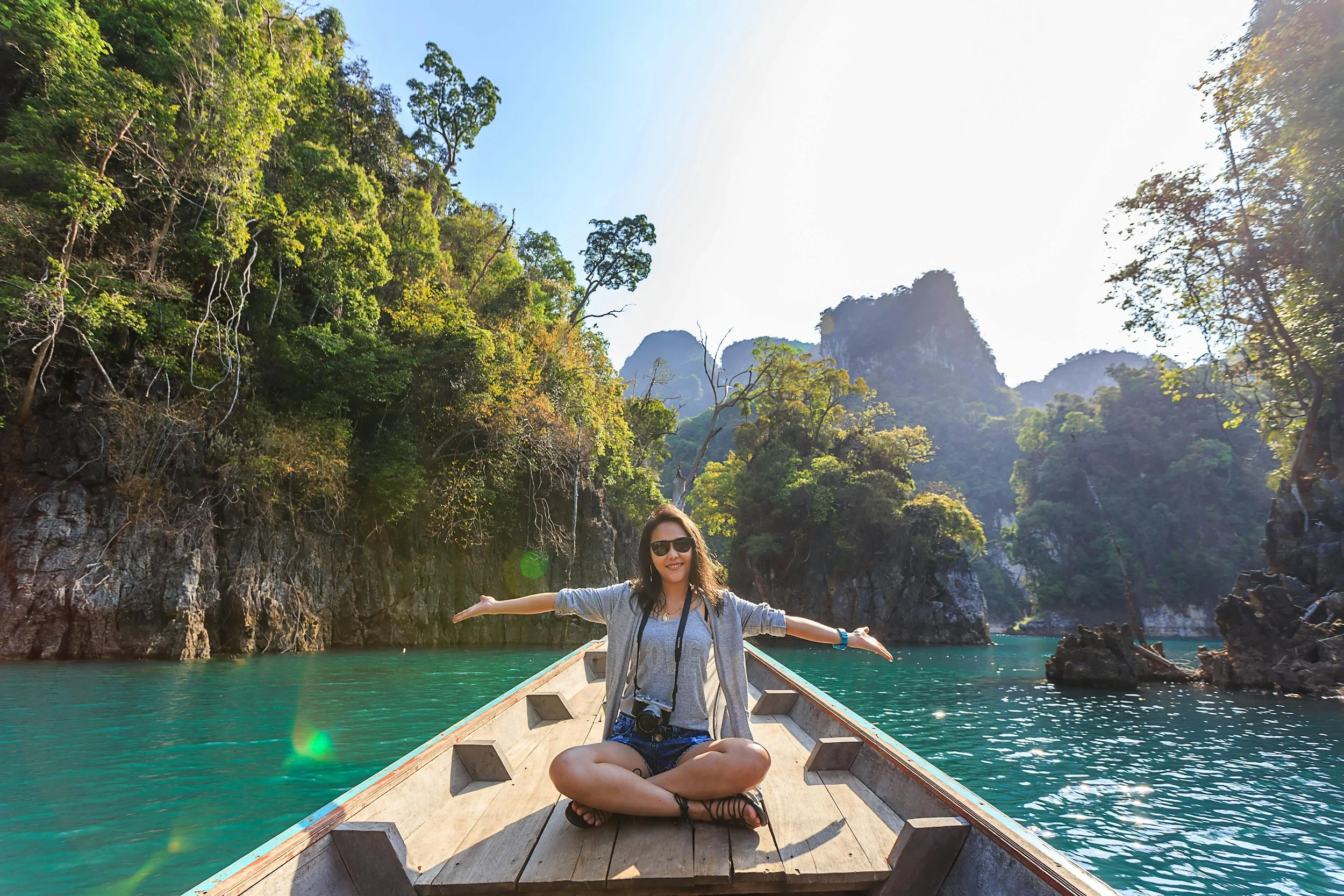 A woman sitting cross-legged on a boat with her arms outstretched, wearing sunglasses, in a lush green river landscape with towering cliffs and trees in the background.
