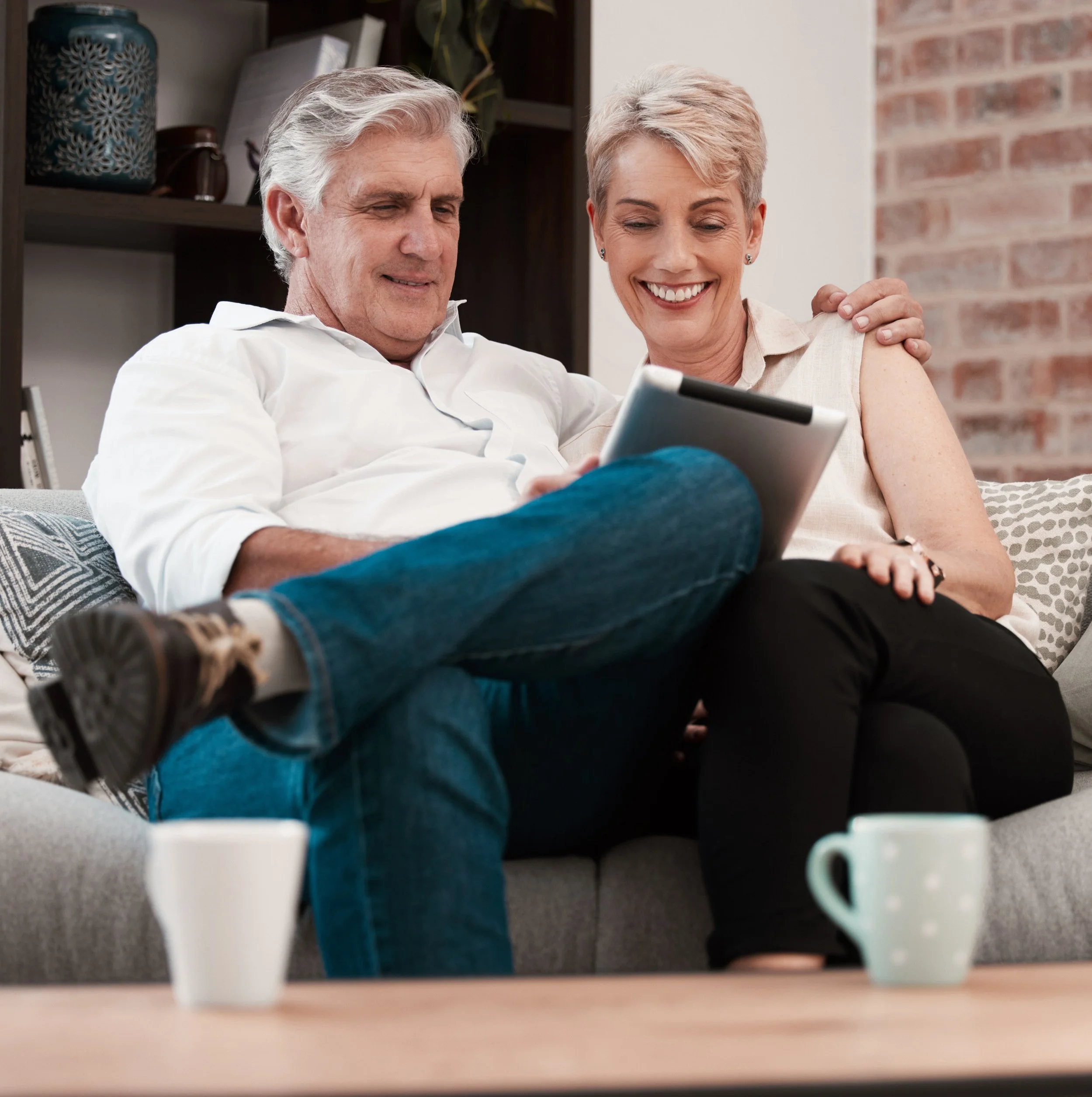 An elderly man and woman sitting on a couch, smiling at a tablet, with coffee mugs on a table in front of them.