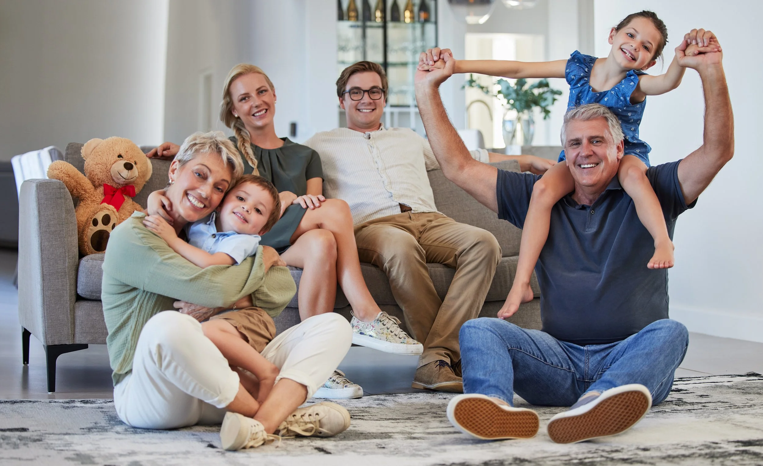 Family of seven, including a man, woman, two girls, two boys, and an elderly man, sitting and playing together on the living room floor and couch, smiling happily in a bright, modern home.