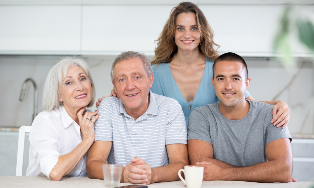 Four family members, two elderly adults, a middle-aged woman, and a young man, smiling and sitting together at a kitchen table.