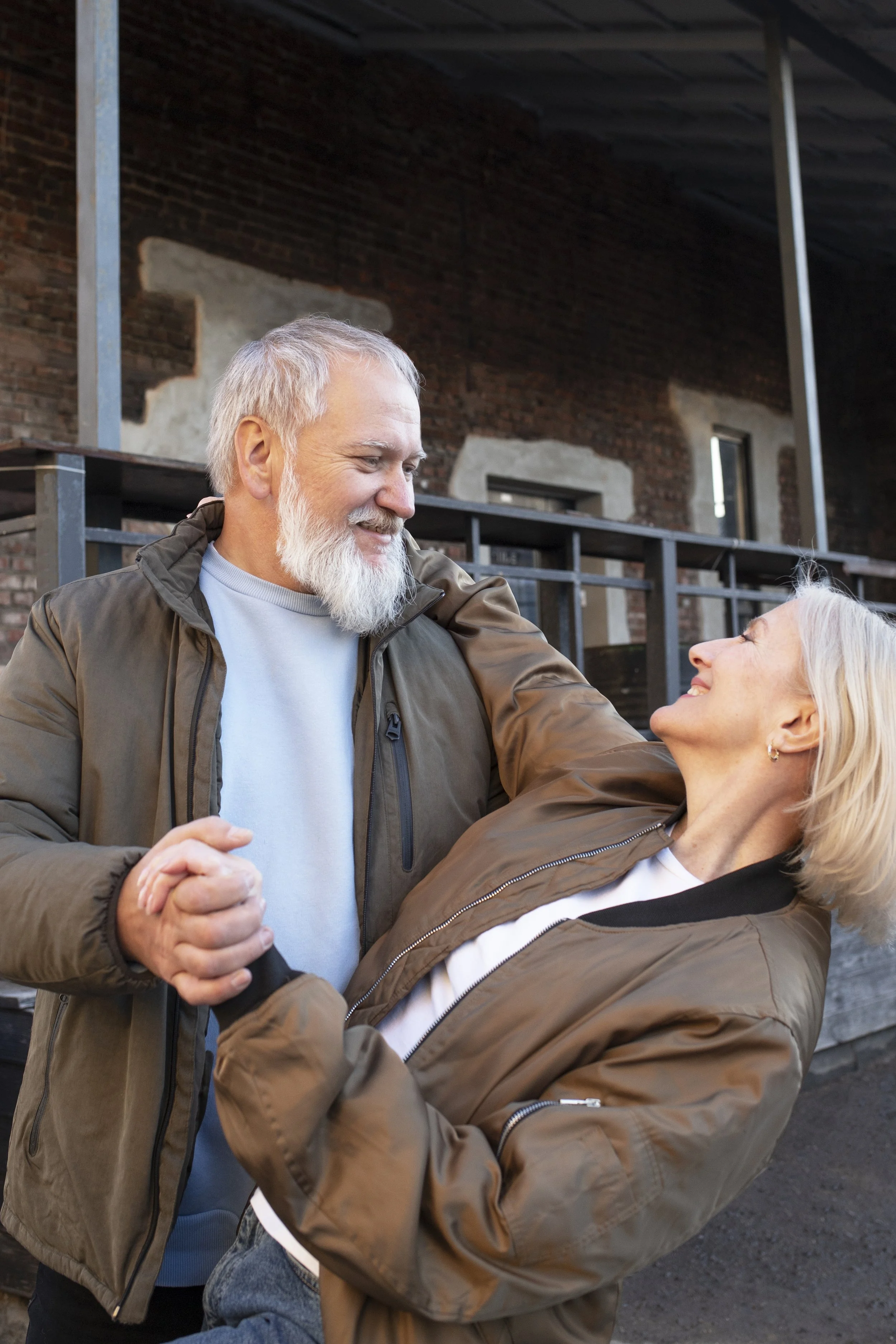 An elderly couple dancing outdoors, the man holding the woman in a dip, both smiling and wearing casual jackets, with a brick building in the background.