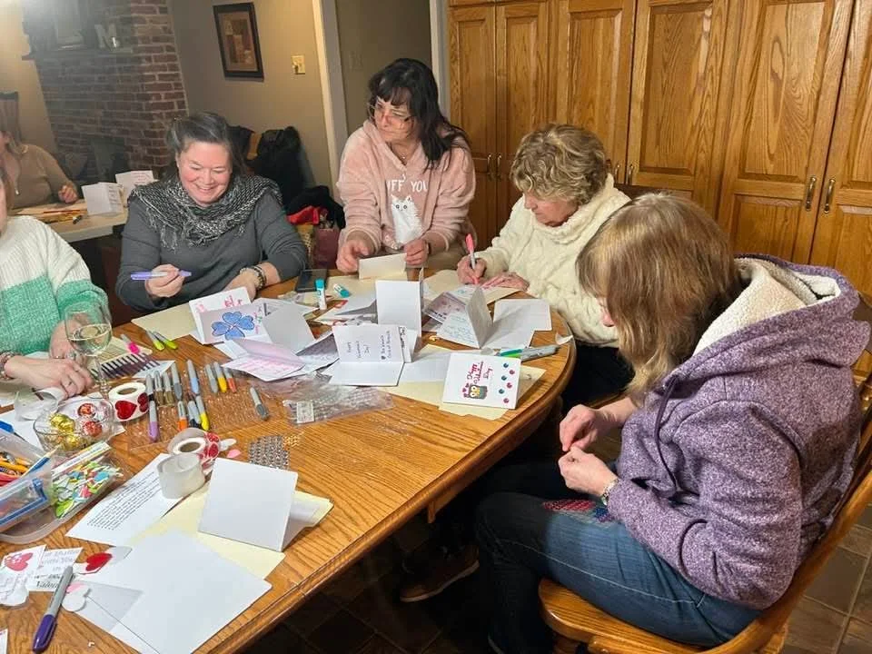 Group of women gathered around a table making greeting cards with various craft supplies in a cozy indoor setting.