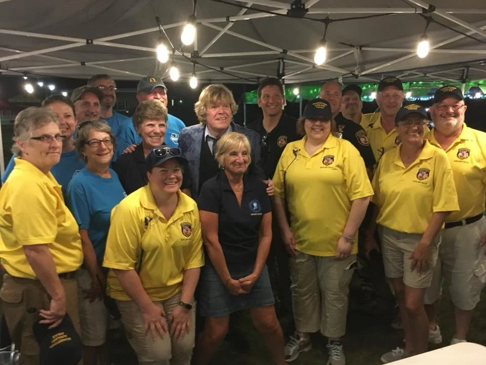 Group of people smiling, some in yellow shirts and hats, under a canopy adorned with string lights at night.