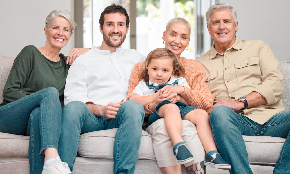 A multi-generational family sitting on a beige sofa in a well-lit room, smiling at the camera.