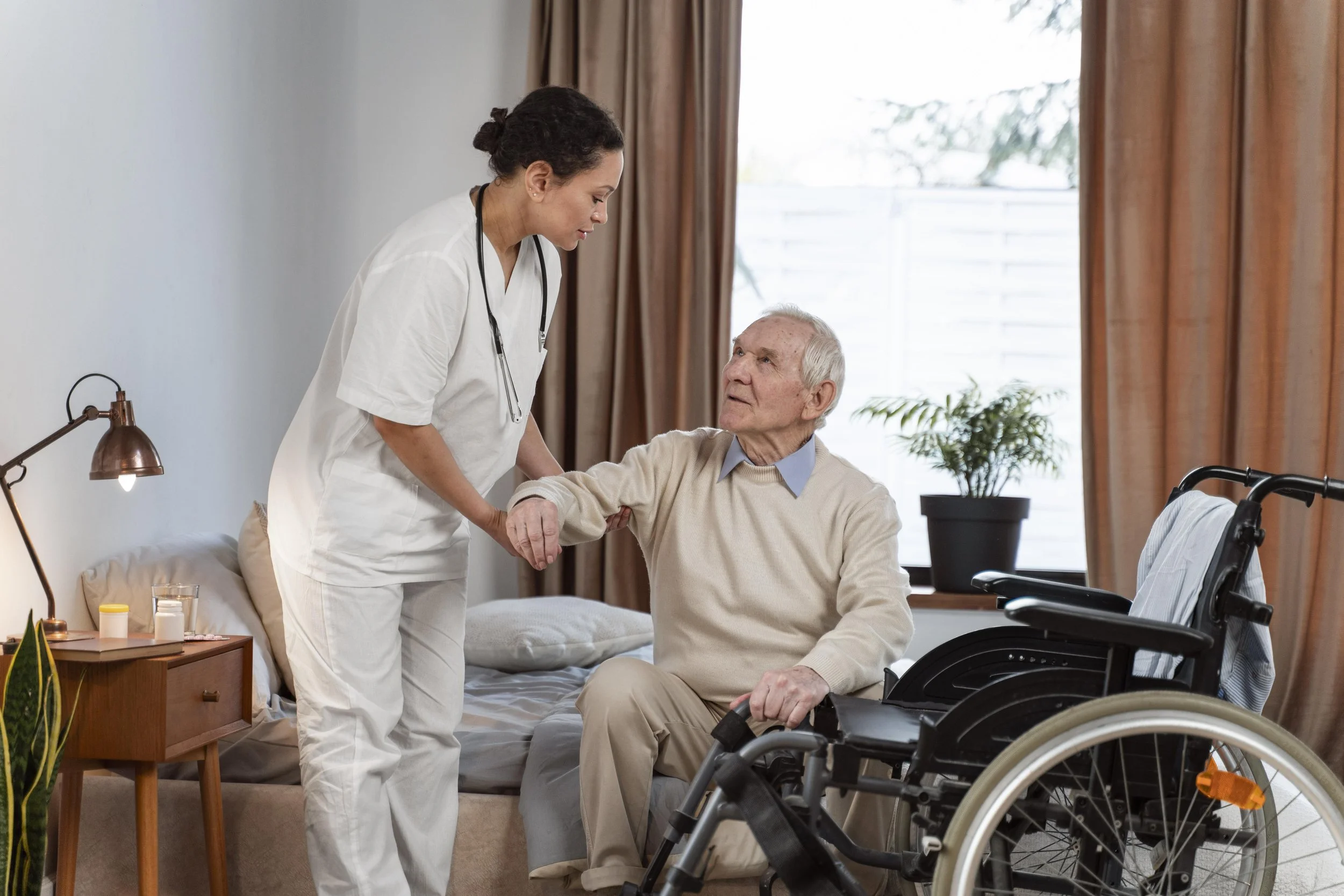 A female nurse helping a senior man in a wheelchair sit on a bed in a cozy bedroom.