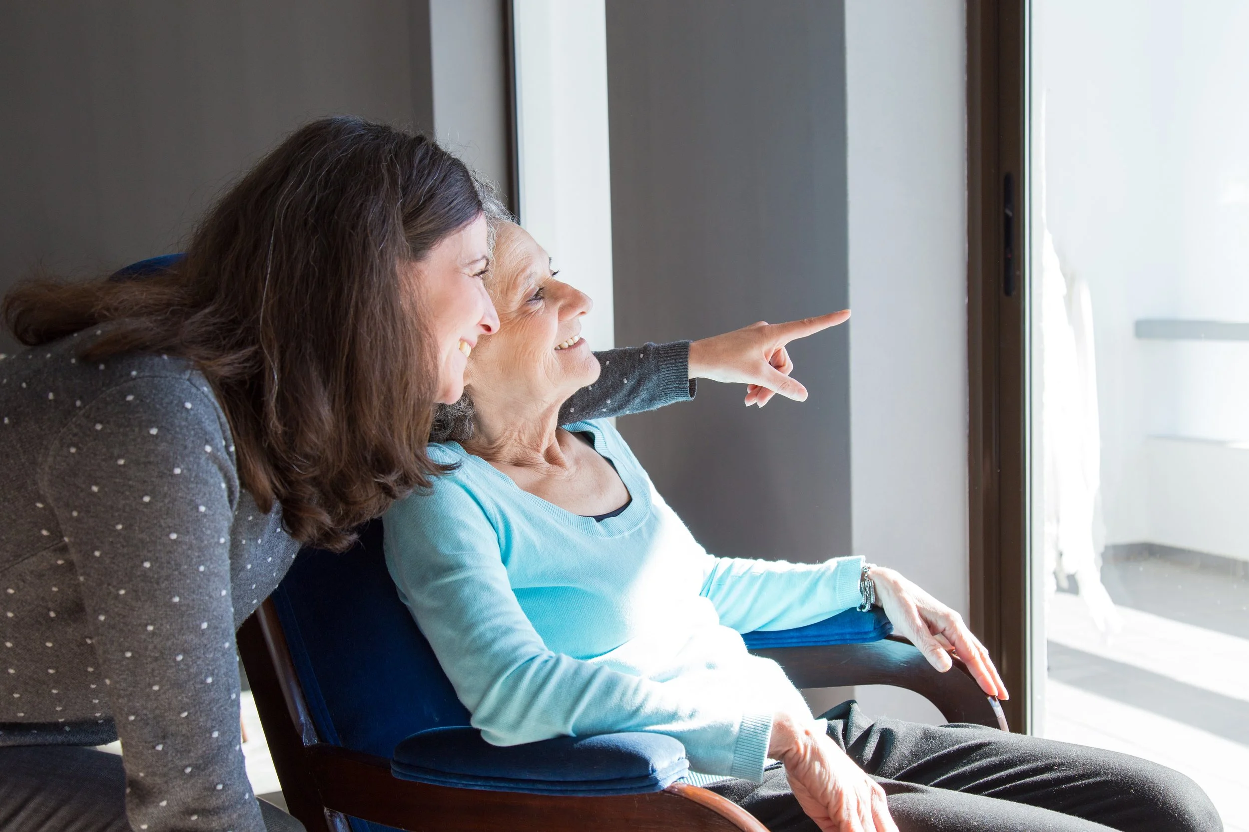 Two women, one elderly and one middle-aged, sitting near a window, smiling and pointing outside.