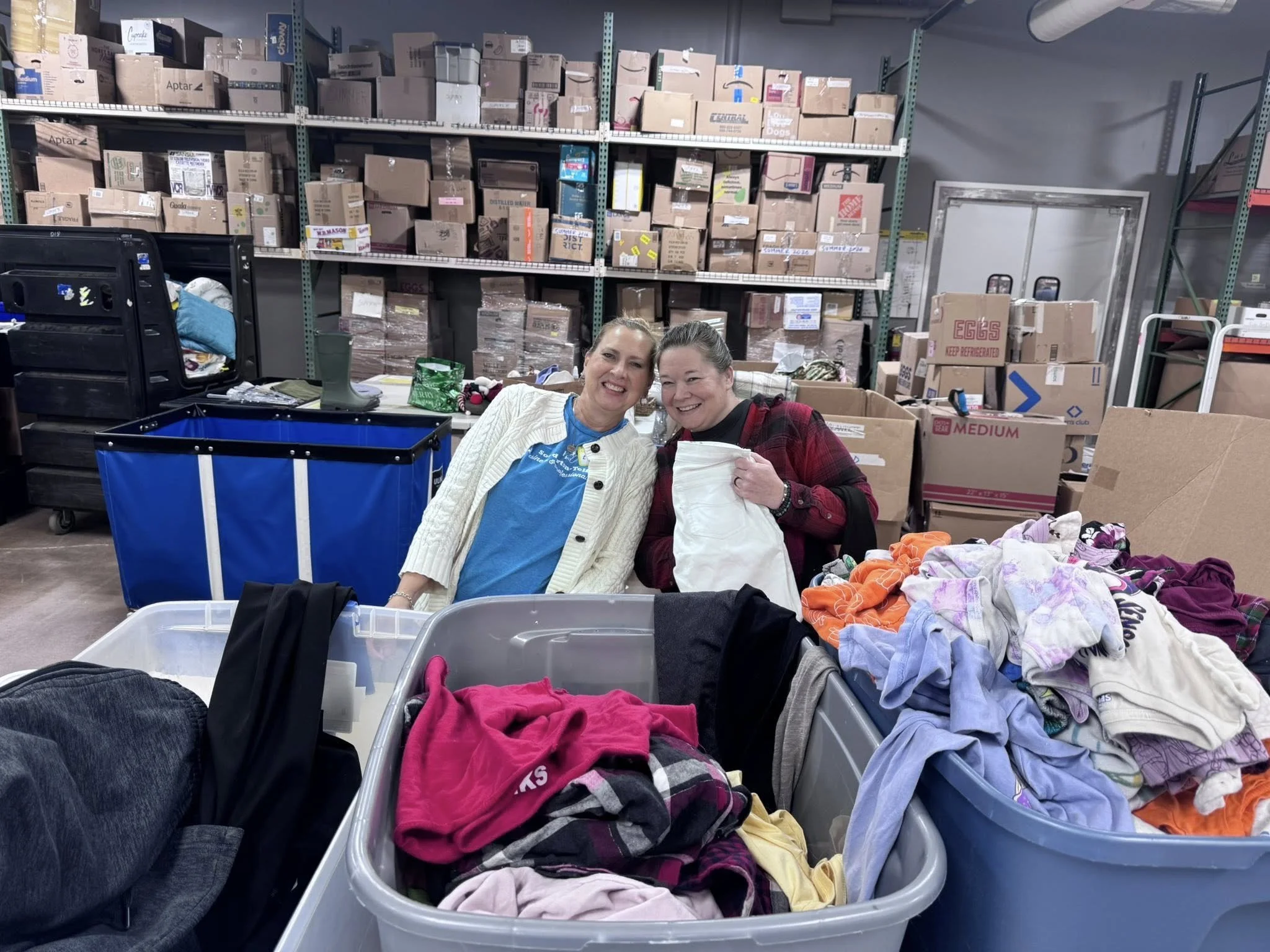 Two women smiling at camera inside a warehouse surrounded by storage bins, cardboard boxes, and clothing, with laundry piles in the foreground.