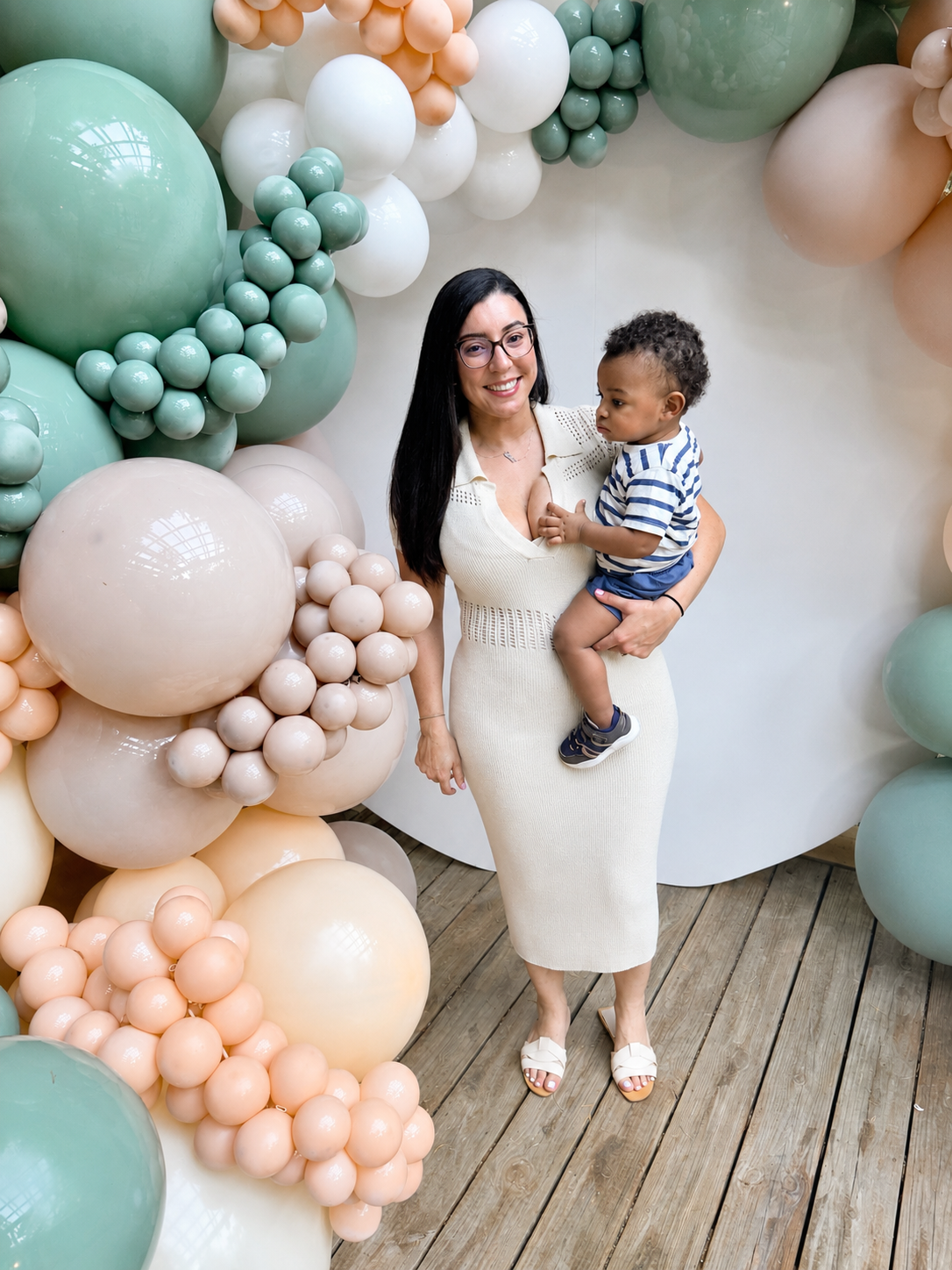 A smiling woman holding a young boy in front of a colorful balloon backdrop at a celebration or event.