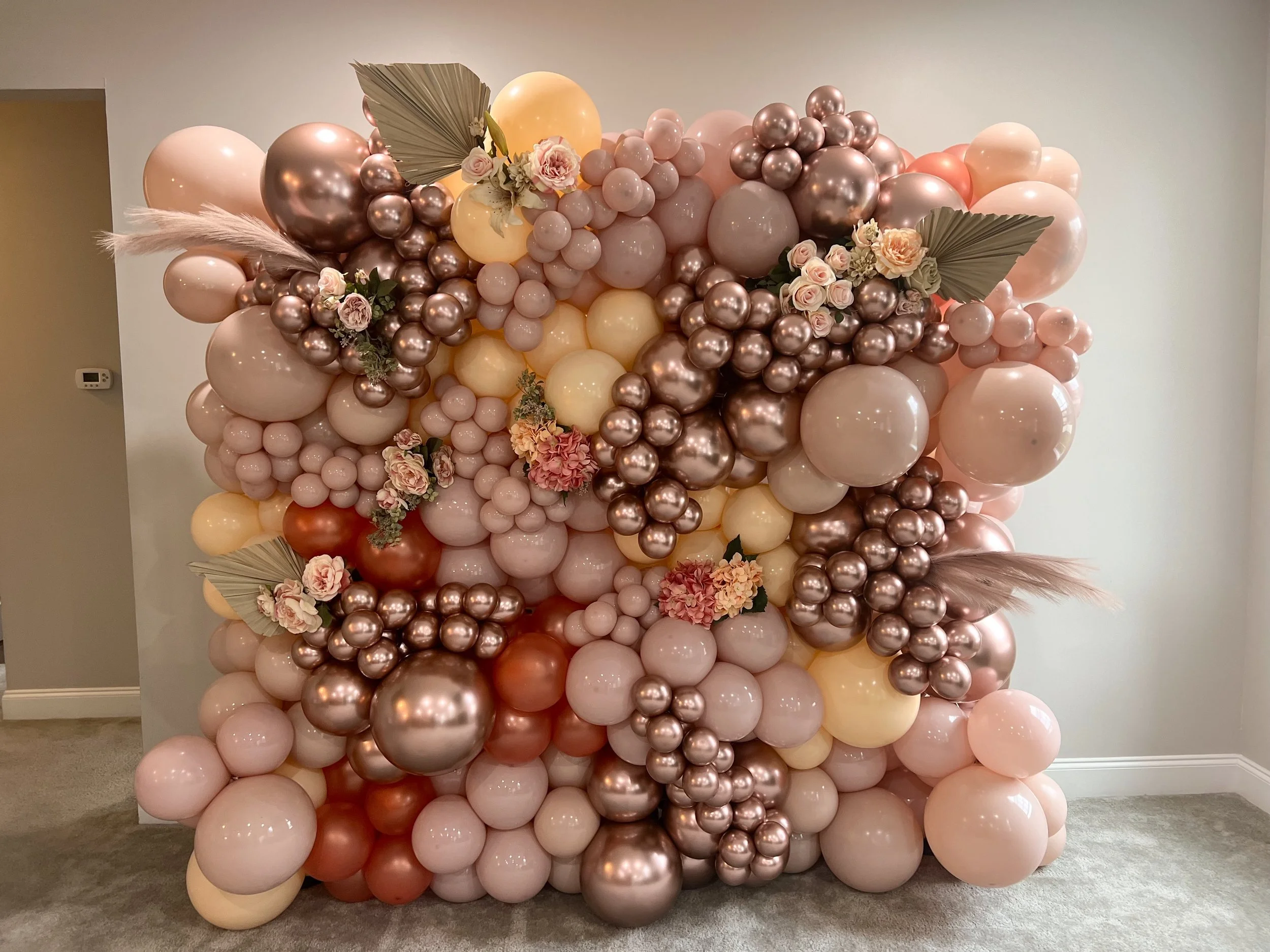 A large balloon arrangement with pink, peach, fertilize, and bronze balloons, decorated with pink roses, hydrangeas, and large dried palm leaves, situated against a plain wall in an indoor setting.
