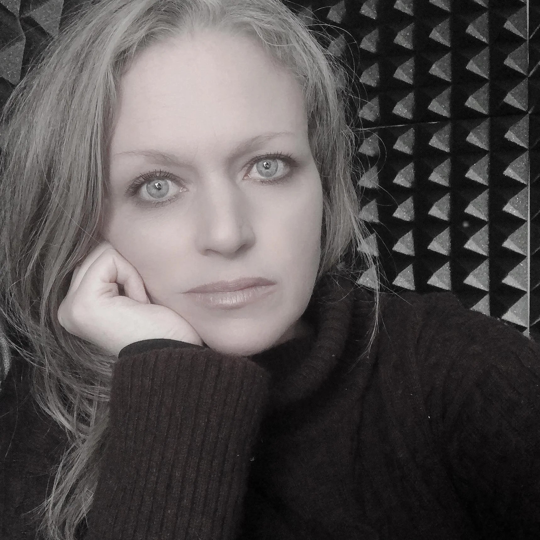 Portrait d'une femme avec des cheveux mouillés, portant un pull noir, reposant sa tête sur sa main, avec un fond acoustique noir et blanc.