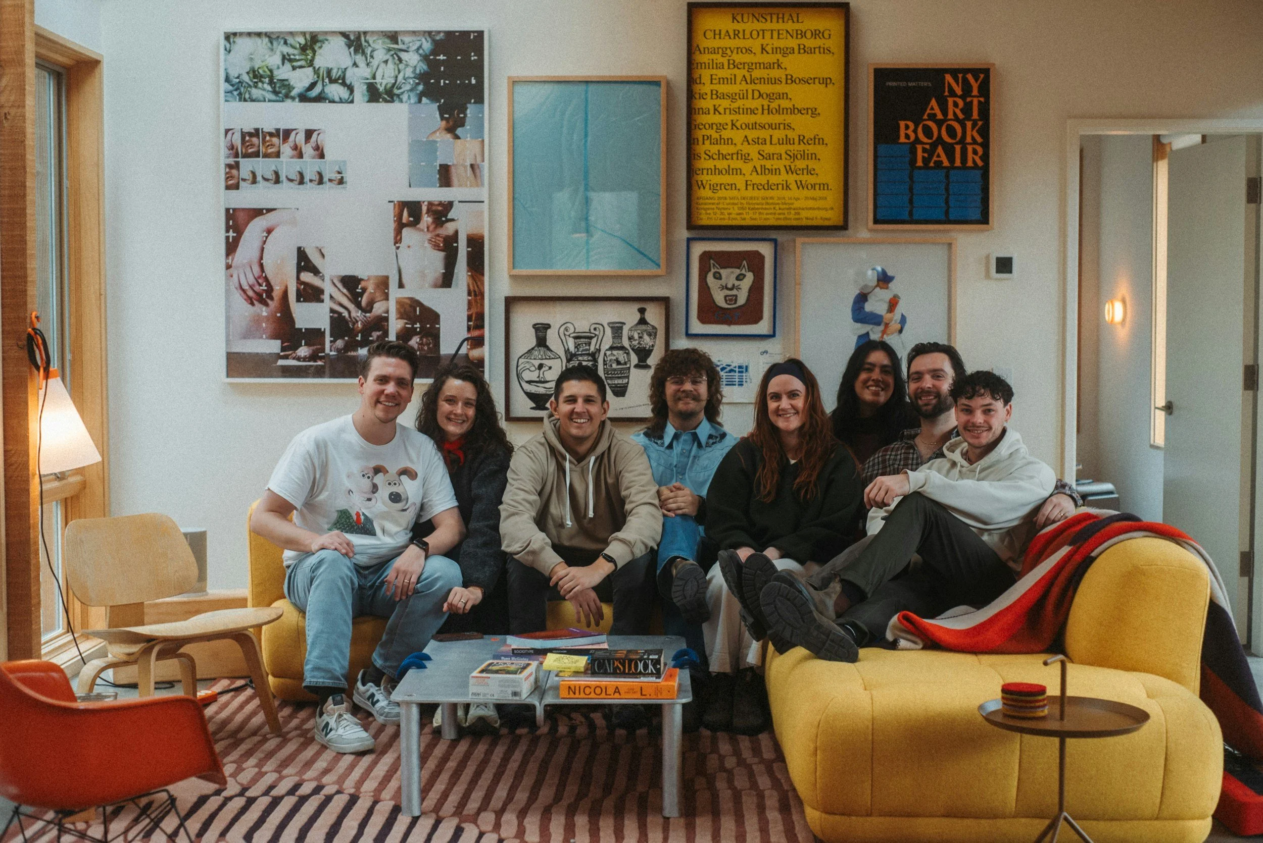 A group of eight people sitting on a yellow couch and a yellow armchair in a living room, smiling at the camera. Behind them are artworks and posters on the wall, a window to the left, and a doorway to the right.
