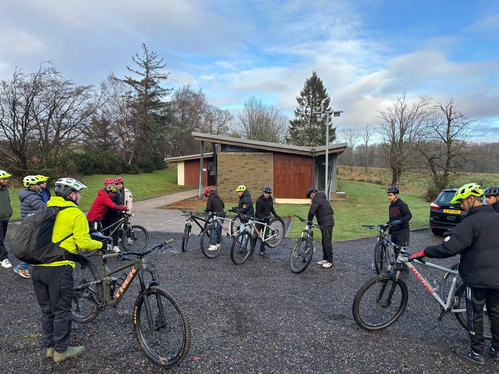 Group of people with mountain bikes gathered outdoors near a modern building on a cloudy day.