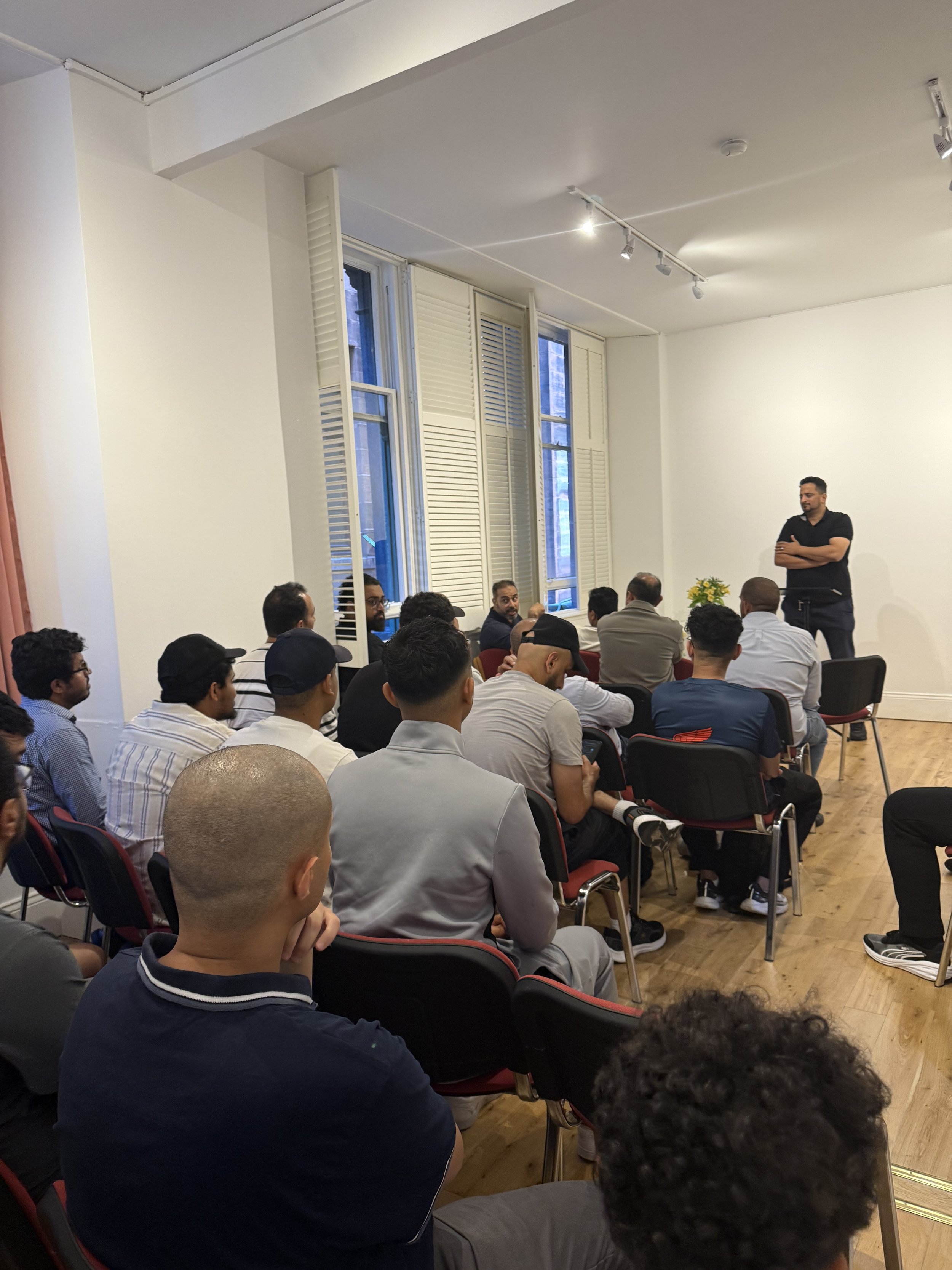 A man standing and speaking to a group of people seated in a room with white walls, wooden flooring, and tall windows with open shutters.