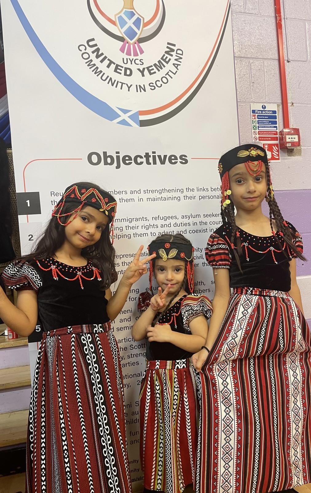 Three young girls wearing traditional Yemeni attire with black tops and red, white, and black patterned skirts and headbands, standing in front of an informational poster about the United Yemen Community in Scotland. الجالية اليمنية في جلاسجو ادنبره