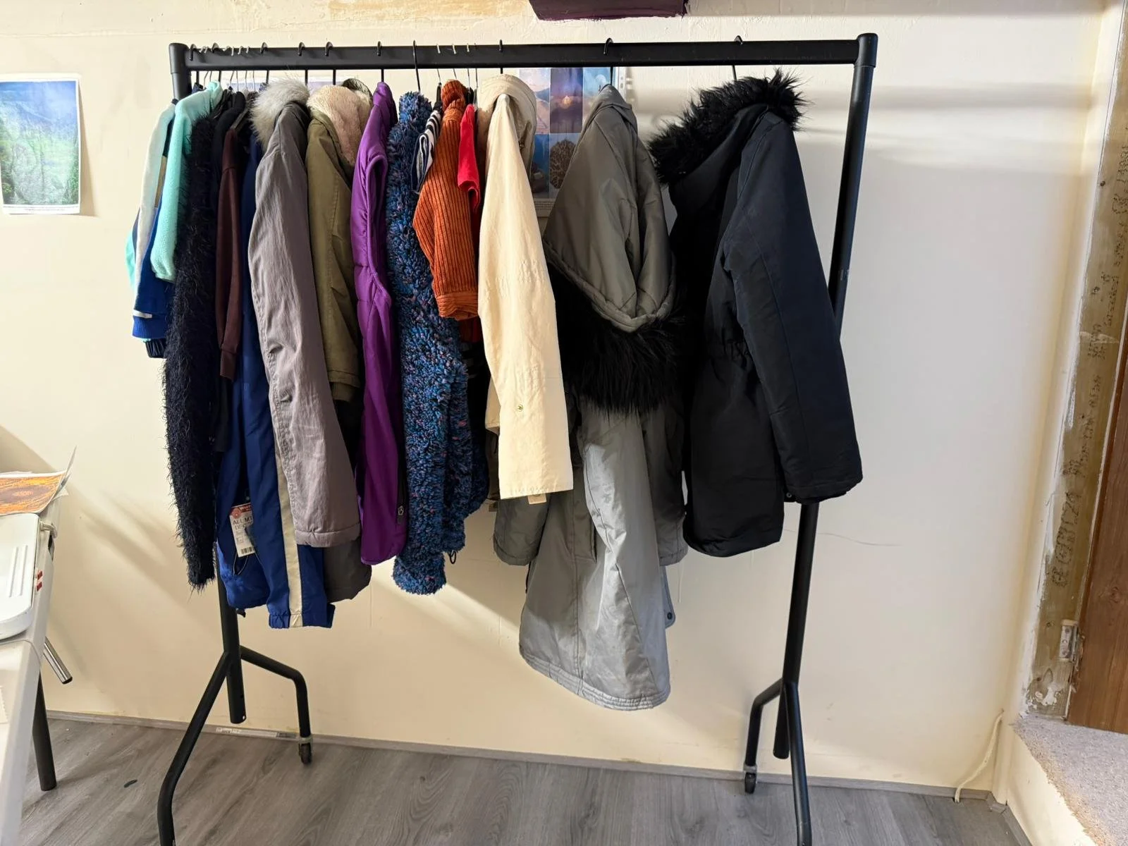 A black clothing rack with various winter jackets and coats hanging on it, placed against a light-colored wall in a room with gray wood flooring.