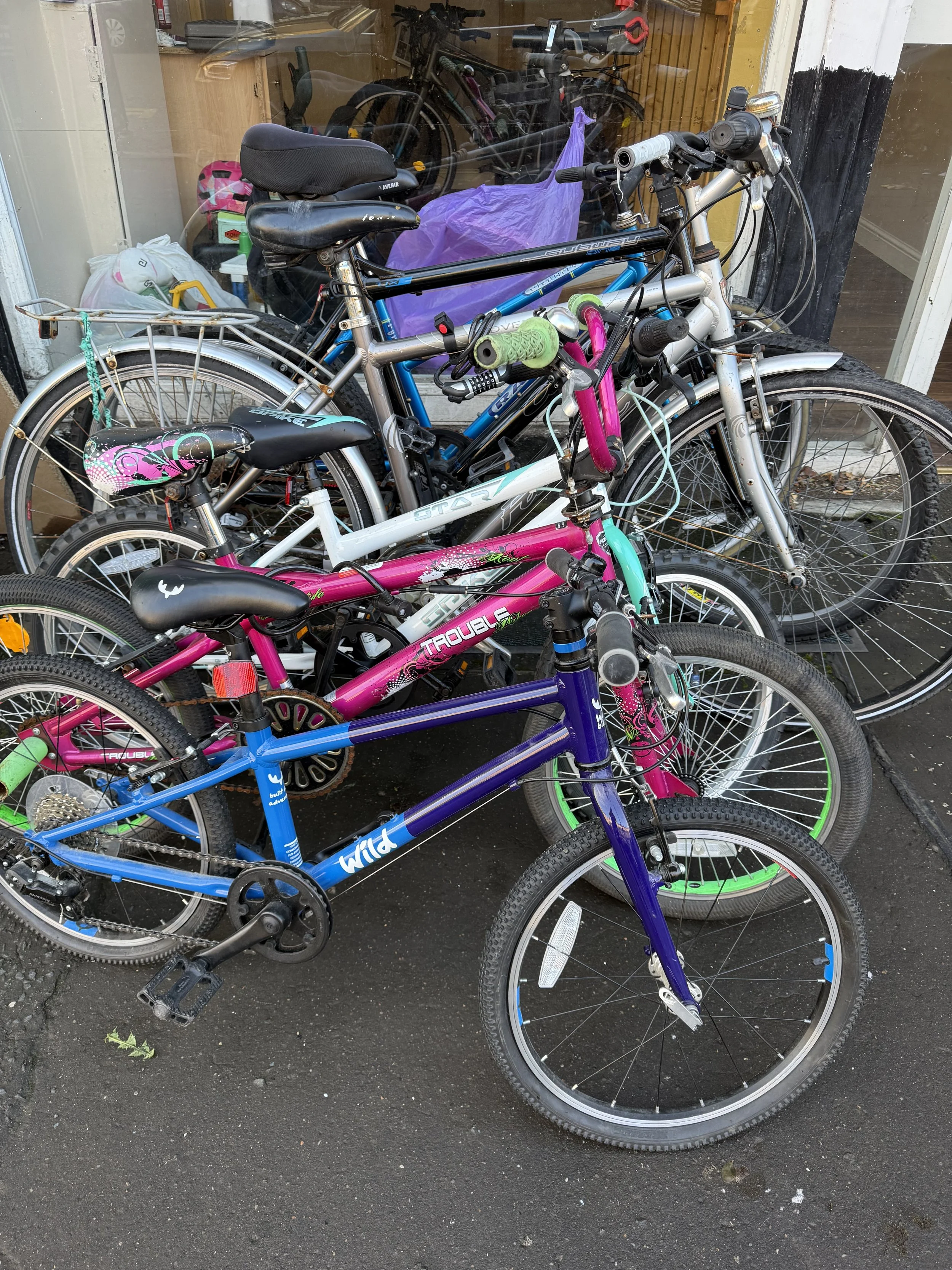 Group of parked bicycles outside a shop, including a blue, purple, pink, and silver bike, some with accessories like a helmet and lights.