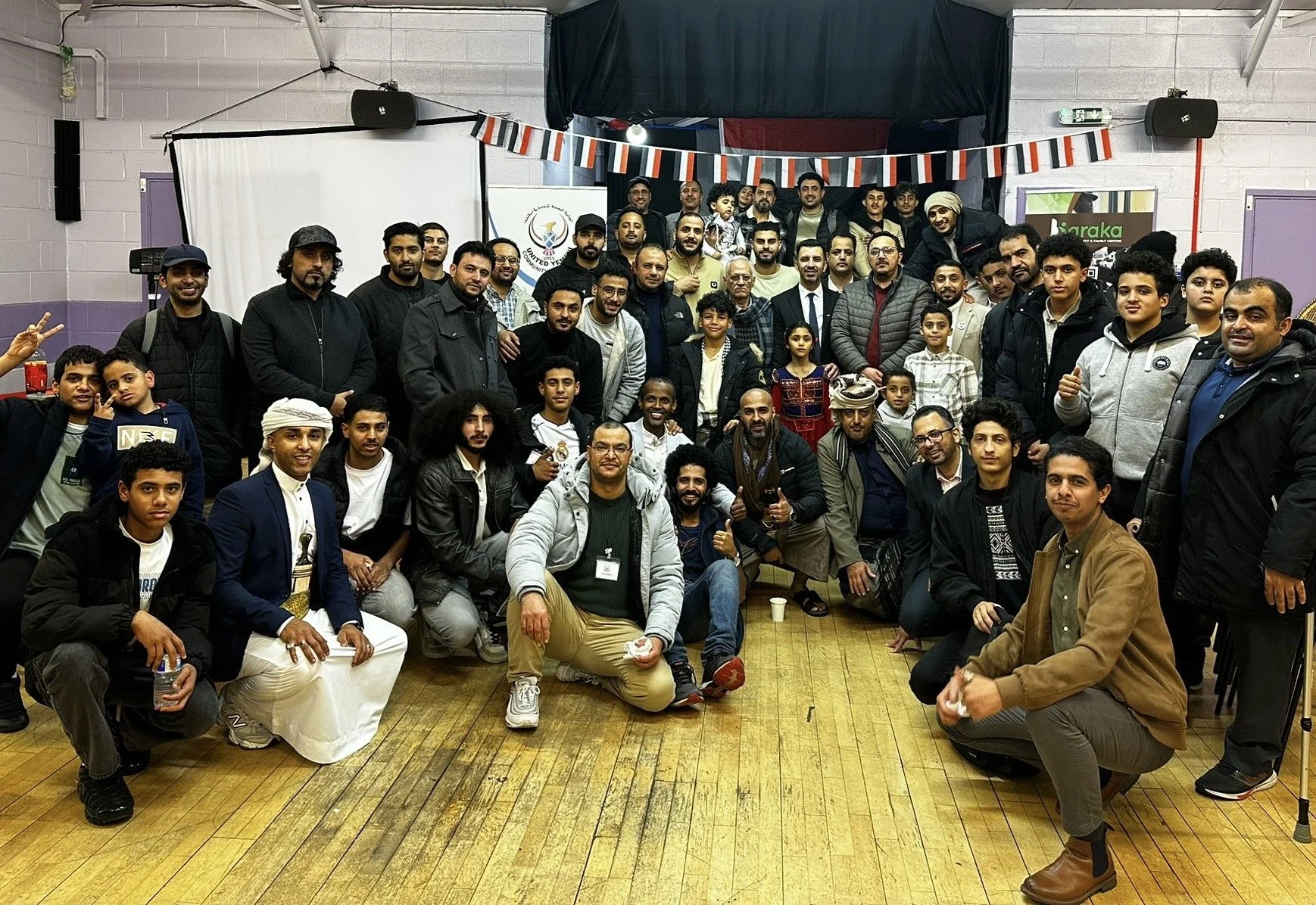 A large group of people posing for a group photo in an indoor setting, with some individuals squatting in the front row and others standing behind. The background features a banner, a projector screen, and some decorations. The group includes men, women, and children of diverse ages and appears to be attending a celebration or gathering.