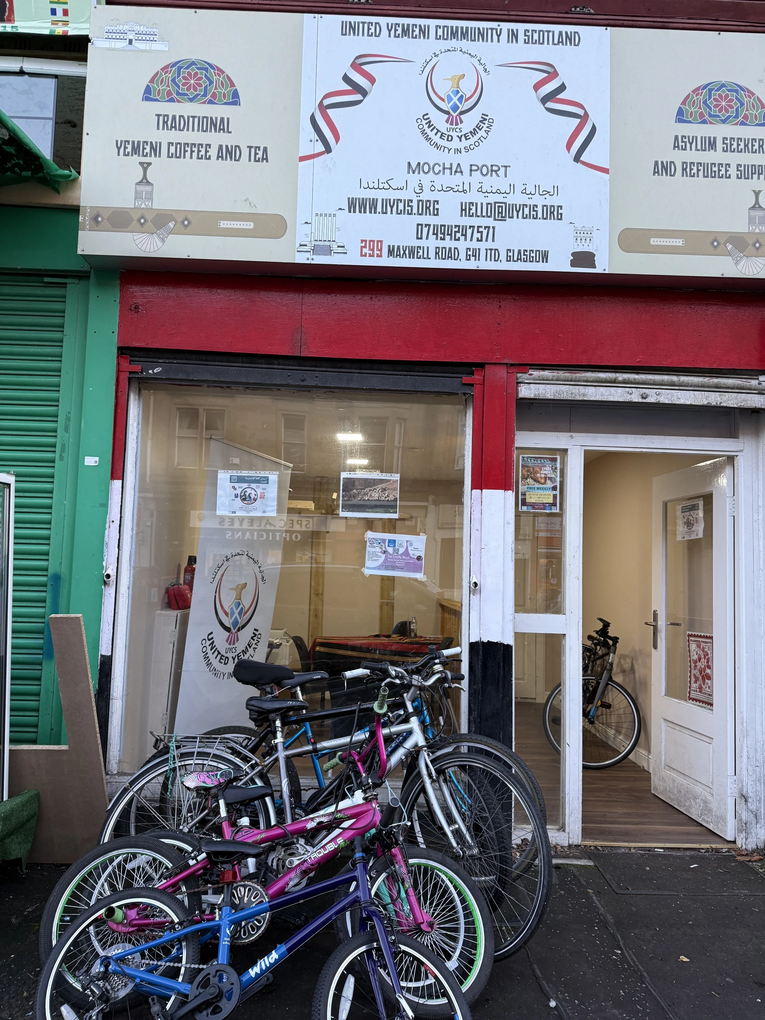 Exterior of a community center with bicycles parked outside. Signage for the United Yemeni Community in Scotland, including information about traditional Yemeni coffee and tea, a mosque, and refugee support programs.
