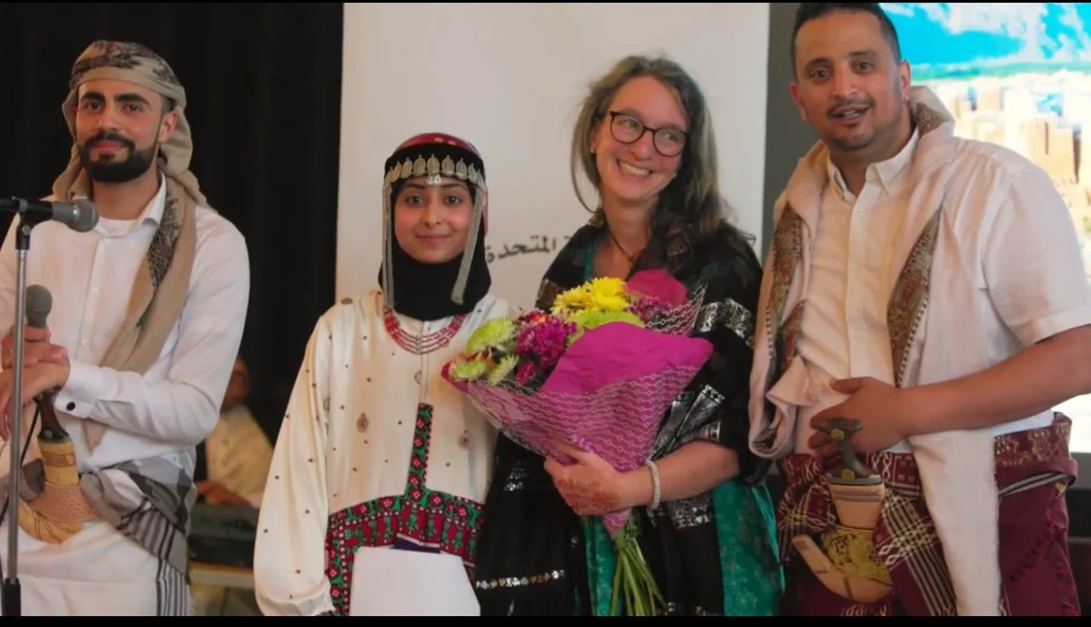 Four people standing together, with one woman holding a bouquet of flowers. The woman and the man on the right are dressed in traditional Middle Eastern attire. The woman in the center is smiling and wearing glasses, with a floral arrangements. A young girl in traditional clothing stands to the left of the woman with flowers, and another man in traditional attire stands on the far left.
