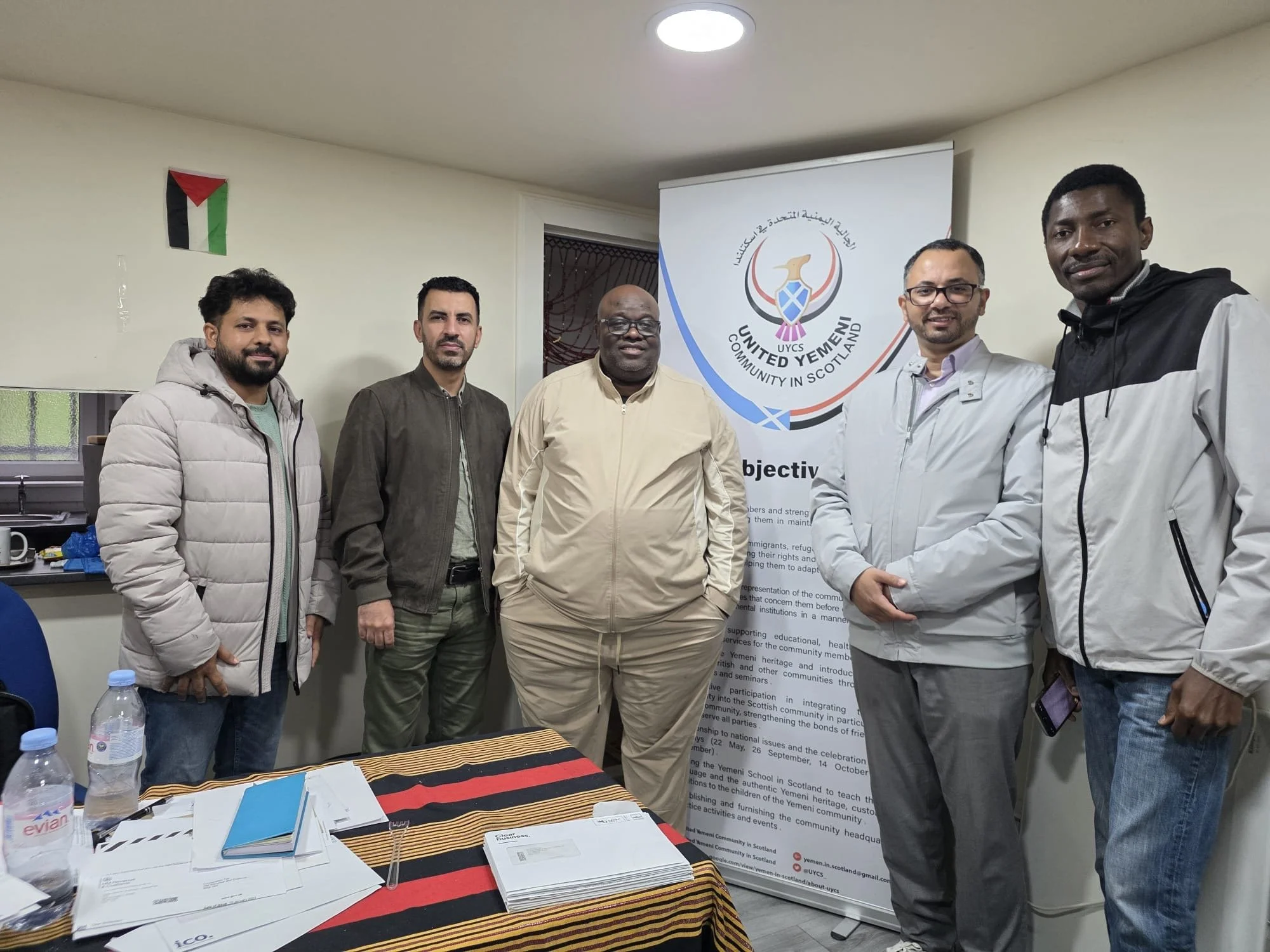 A group of six men standing indoors in front of a United Yemen community banner, with a small Yemeni flag on the wall behind them. They are smiling and dressed in casual jackets.